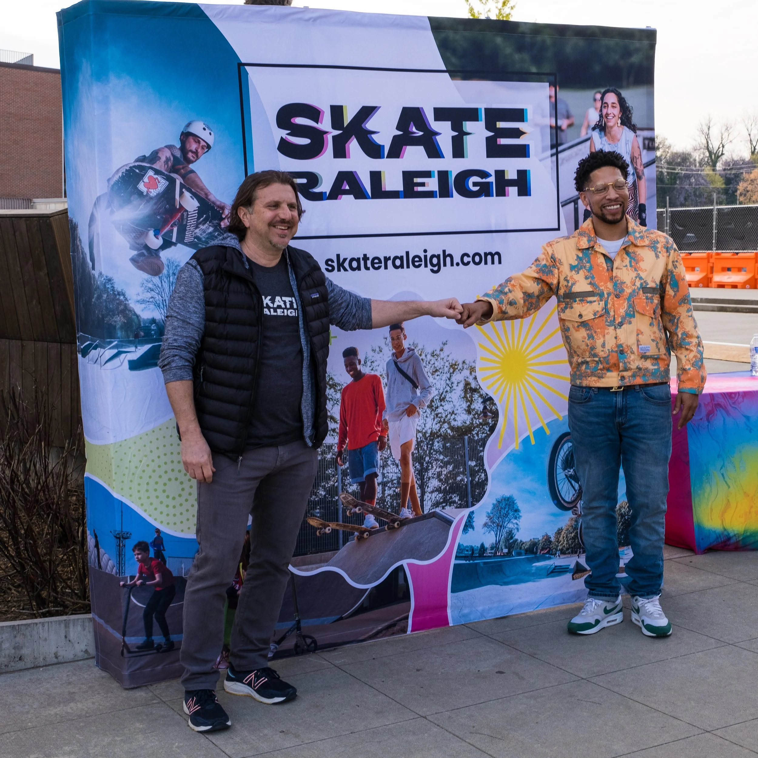 Two men shaking hands in front of a colorful banner at a fundraiser event for for Skater Raleigh.