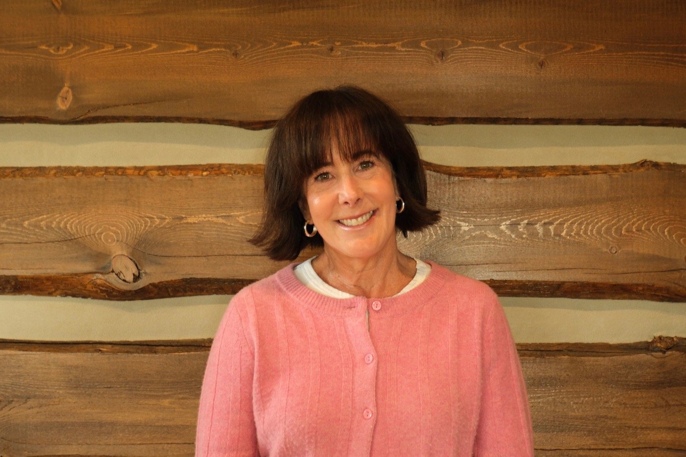 A woman with short dark hair wearing a pink sweater smiling in front of a stacked log wall