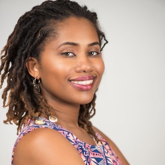 A woman with shoulder length locs wearing a patterned top smiling in front of a neutral background