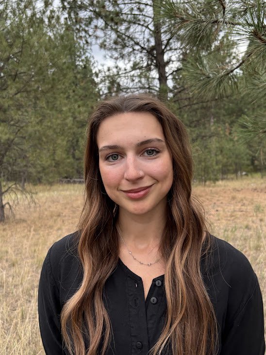 A young woman with long brown hair smiling outside, with pine trees in the background