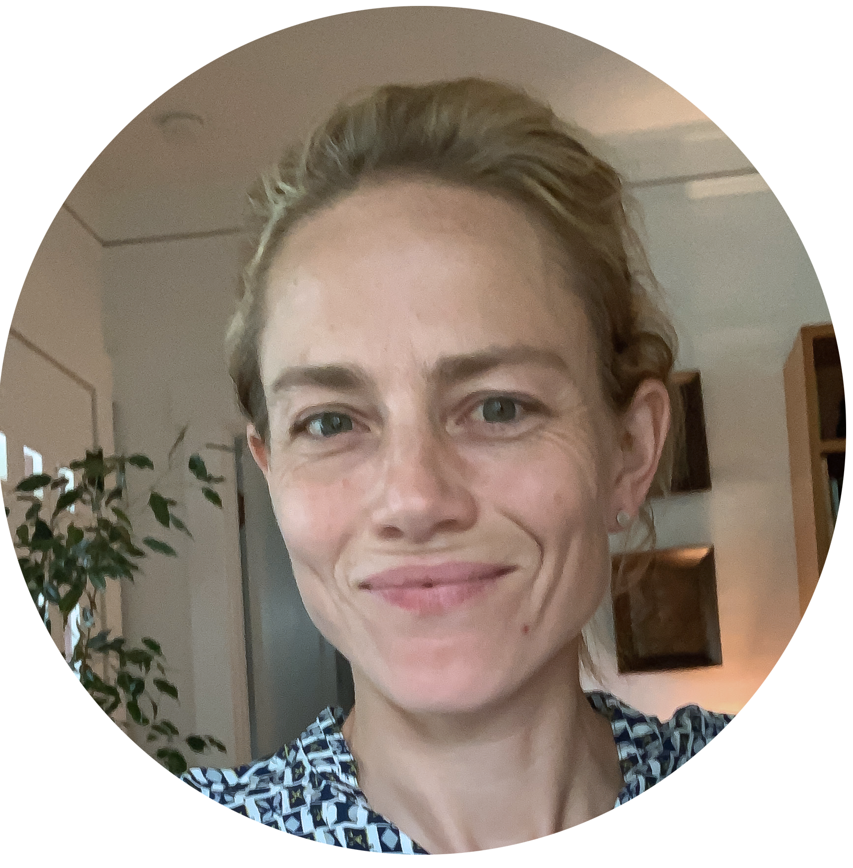 A woman with strawberry blonde hair smiling in a living room, with a plant and bookshelves in the background