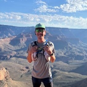 A young man wearing a runner's backpack smiling, standing at the brim of a vast canyon with blue skies above