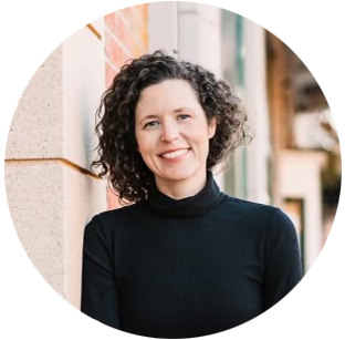 A woman with curly dark hair smiling, wearing a black turtleneck, leaning against a brick wall outdoors
