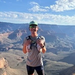 A young man wearing a runner's backpack smiling, standing at the brim of a vast canyon with blue skies above