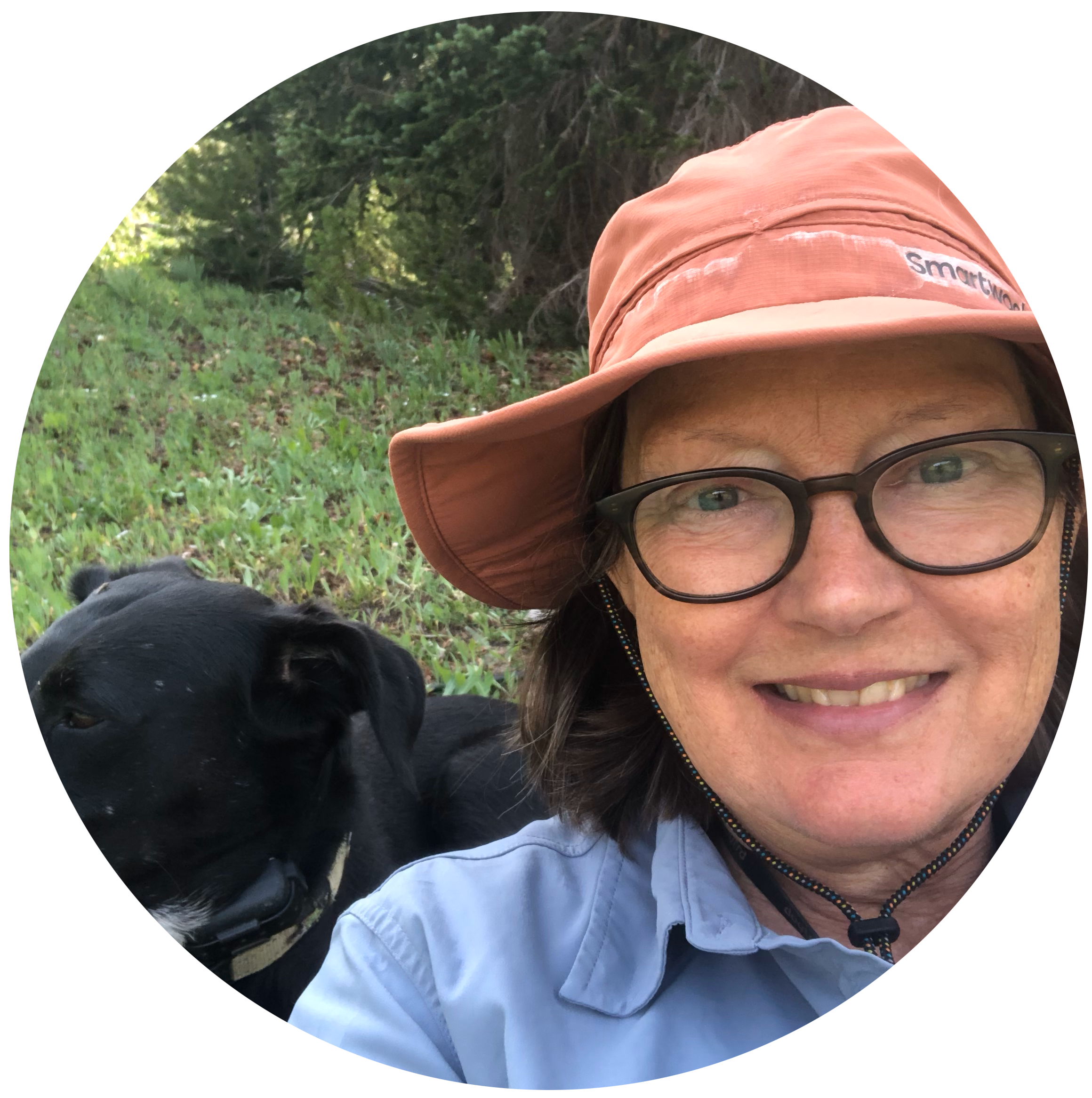 A woman with glasses and a wide-brimmed hat smiling outdoors, with a black dog lying next to her on green grass, trees in the background