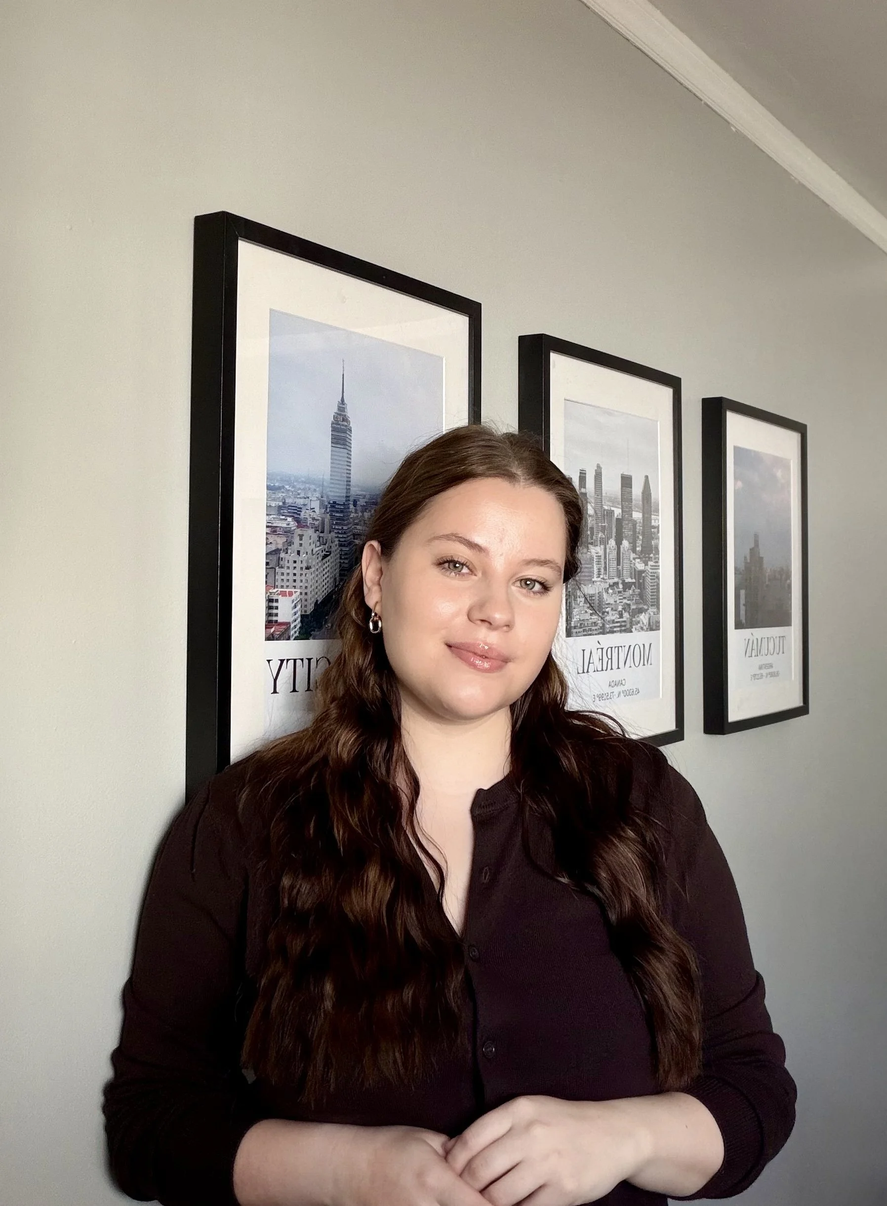 A young woman with wavy brown hair, light skin, and a nose piercing, smiling, dressed in black, with tattoos visible on her arm, against a plain gray background.