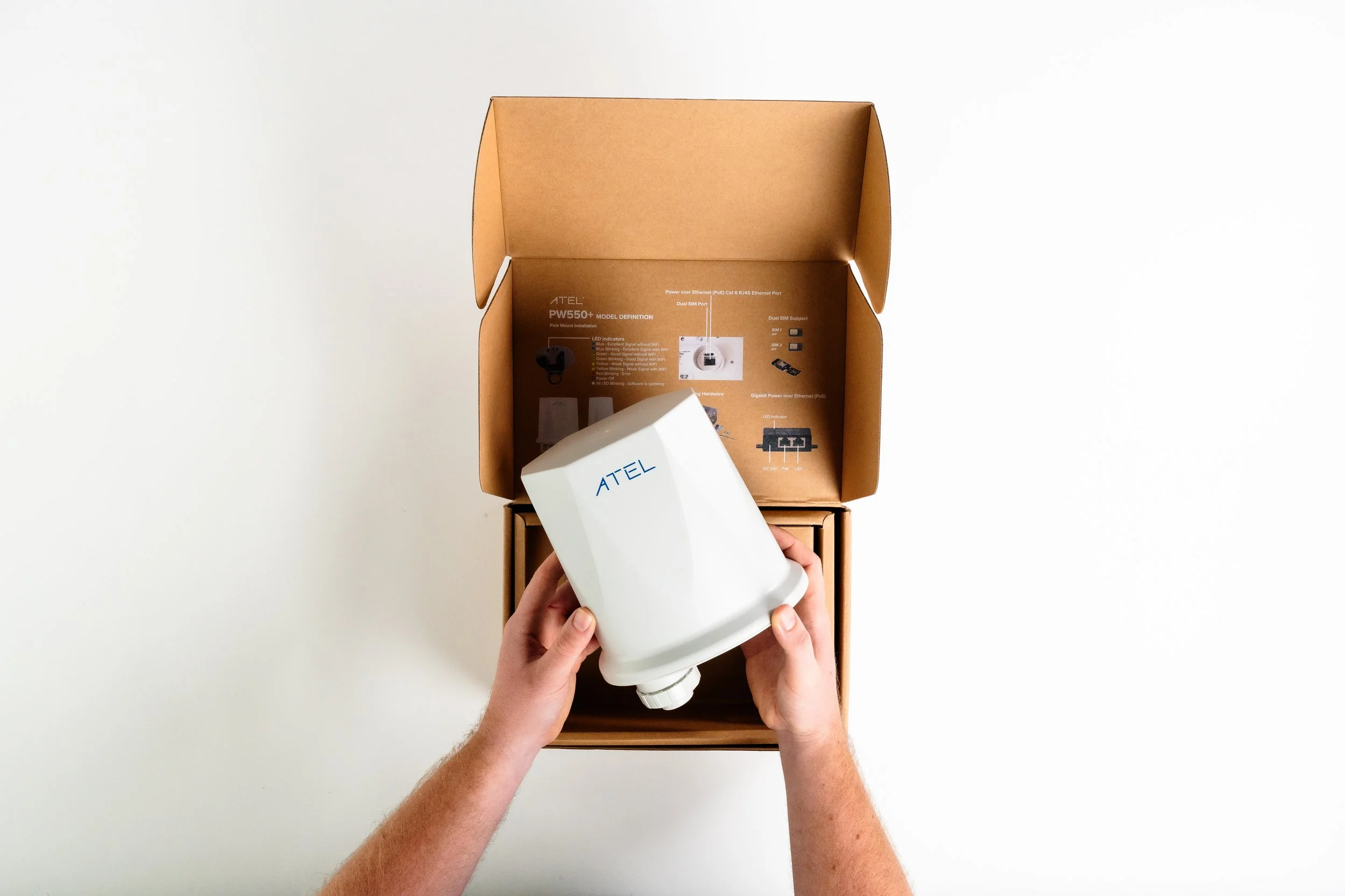 Person holding a white ATEL device over an open cardboard box with product information inside, on a white surface.