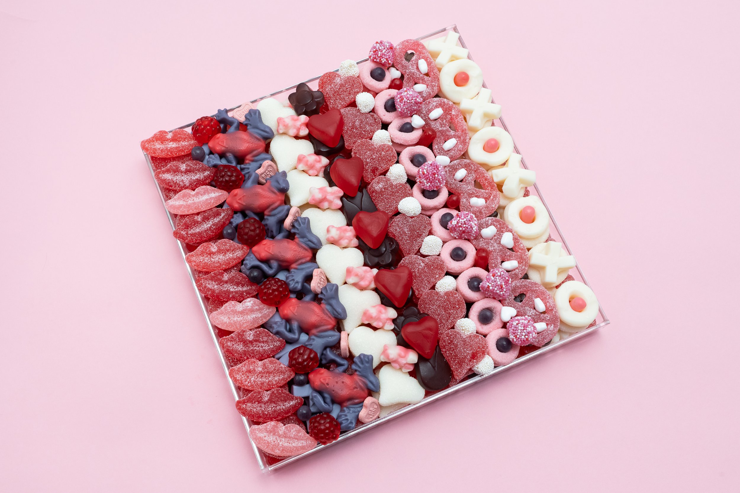Assorted colorful candies on a square glass tray on a pink background.