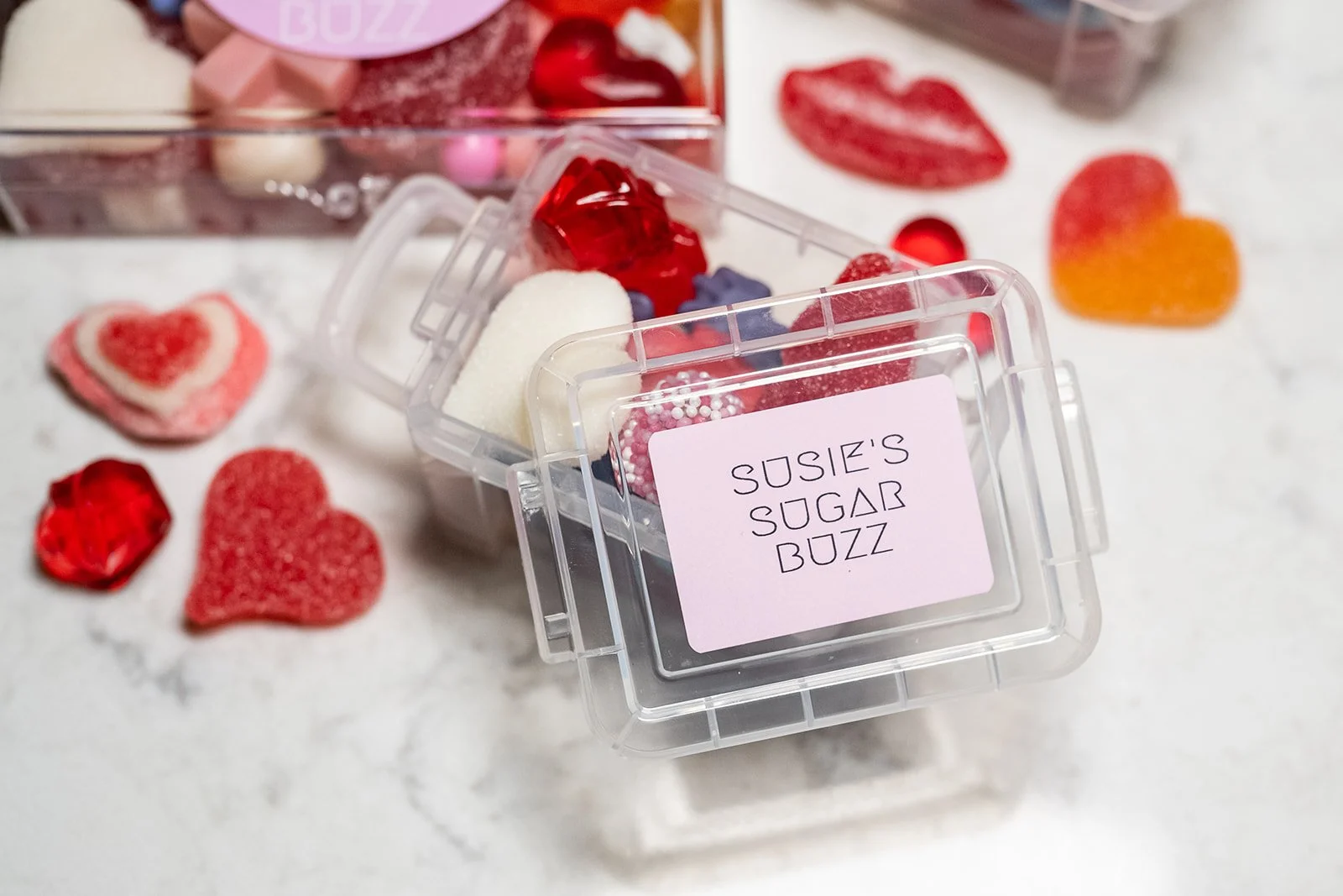 A clear plastic container labeled "Susie's Sugar Buzz" filled with assorted candies, with various heart-shaped candies and gummy candies scattered around on a white surface.