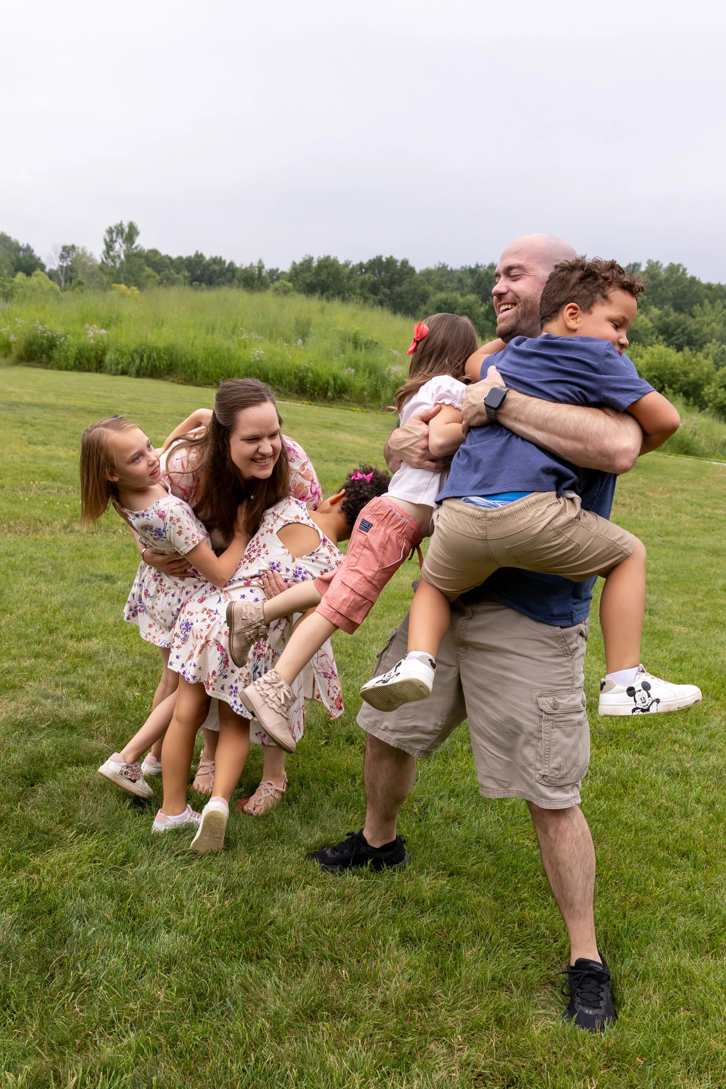 Minneapolis Family Photography - A blended family of a mom, dad and four kids are at Lebanon Hills Regional Park in Eagan, MN. The mom and dad are standing by each other as their four kids jump into their arms all laughing together.