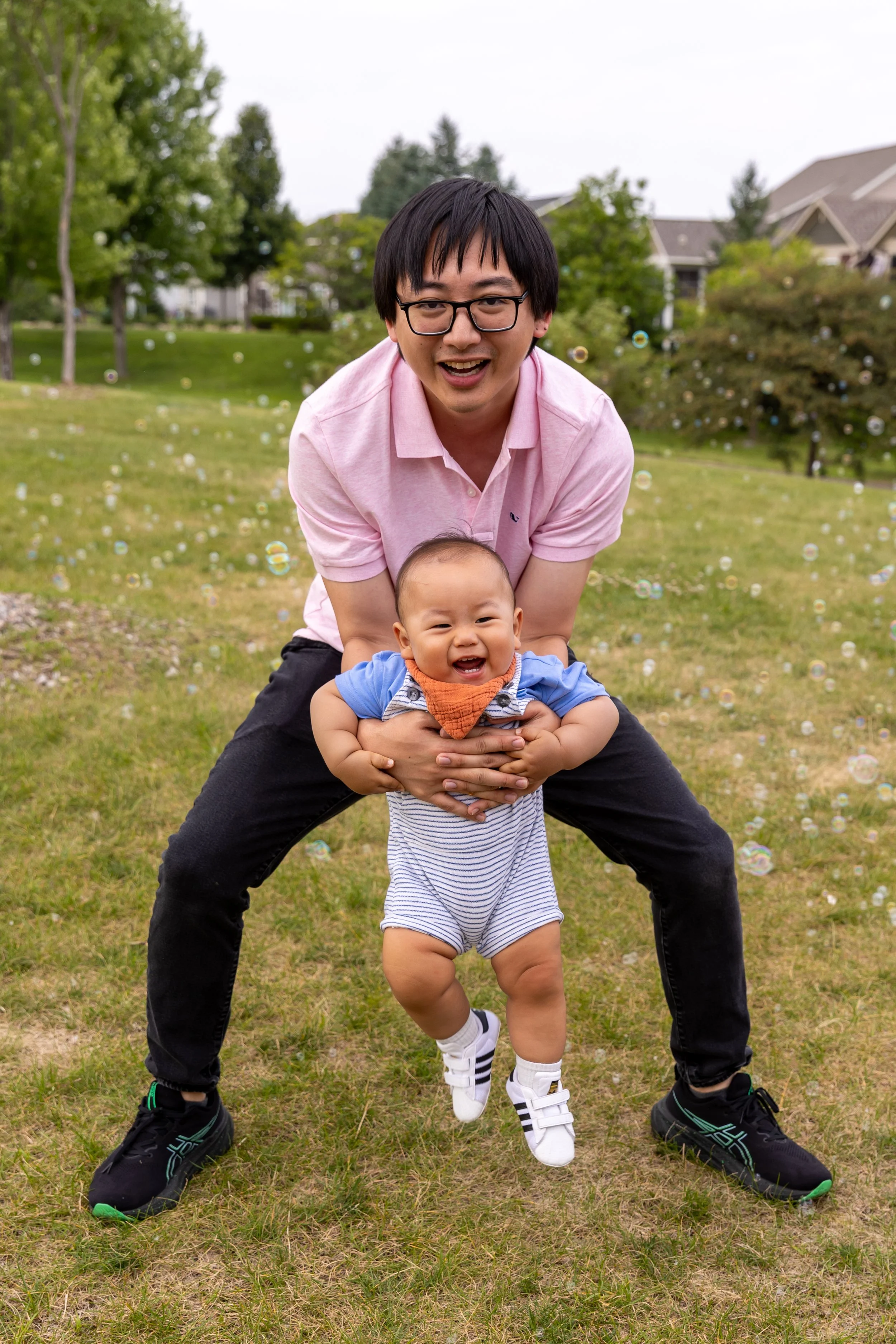 A dad in a pink shirt and black pants holds his son wearing  white and blue striped overalls between his legs as they both smile at the camera in a St. Paul Park.