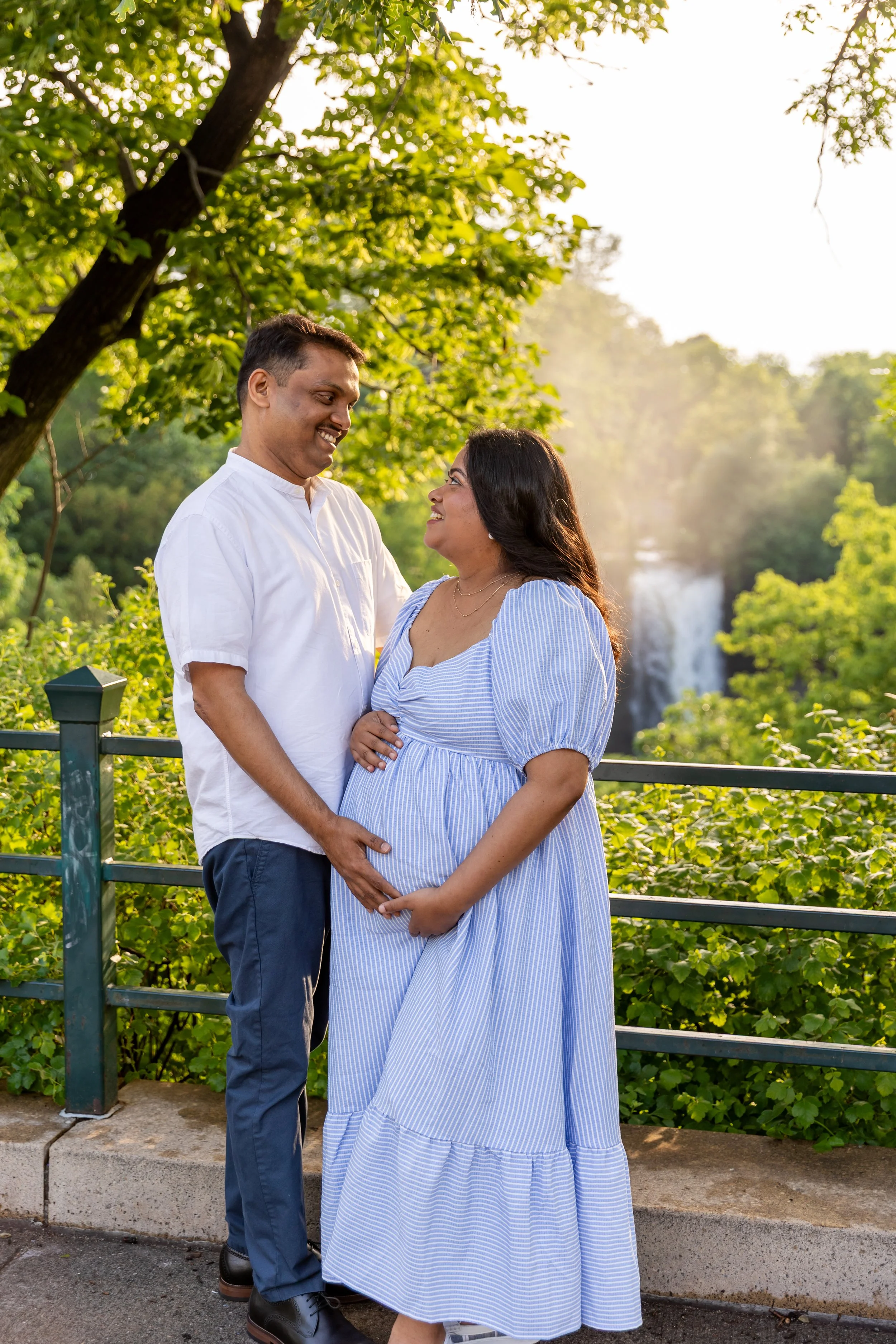Maternity session - father and mother stand together smiling at the camera while cradling each other and the baby bump while at Minnehaha Falls Park with the waterfall in the background