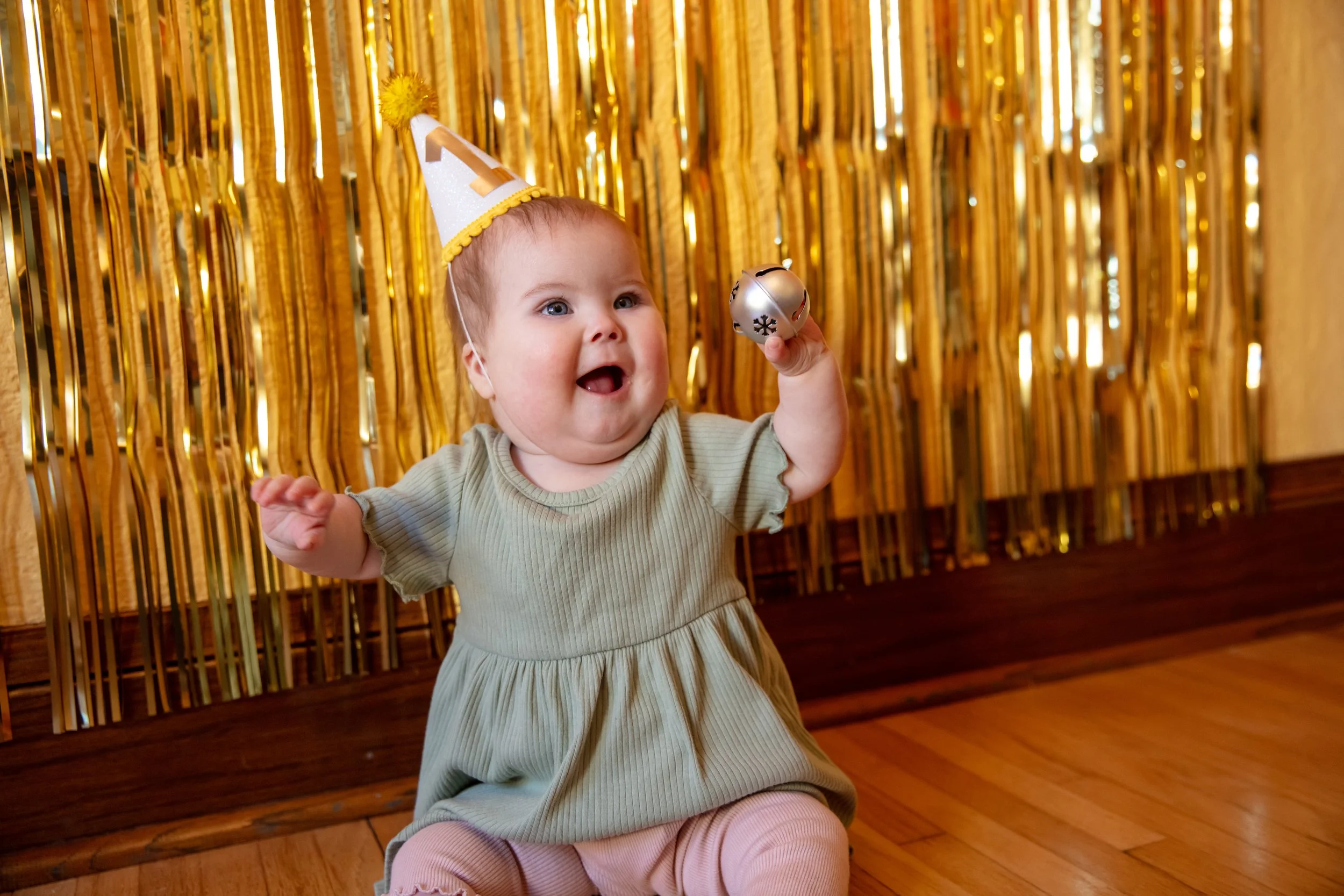 Minneapolis Cake Smash photographer - photo of a one year old baby is sitting on the floor looking up at the camera and is in front of birthday streamers.  She is wearing a green dress and a birthday hat with the number 1 on it and holding a bell in 