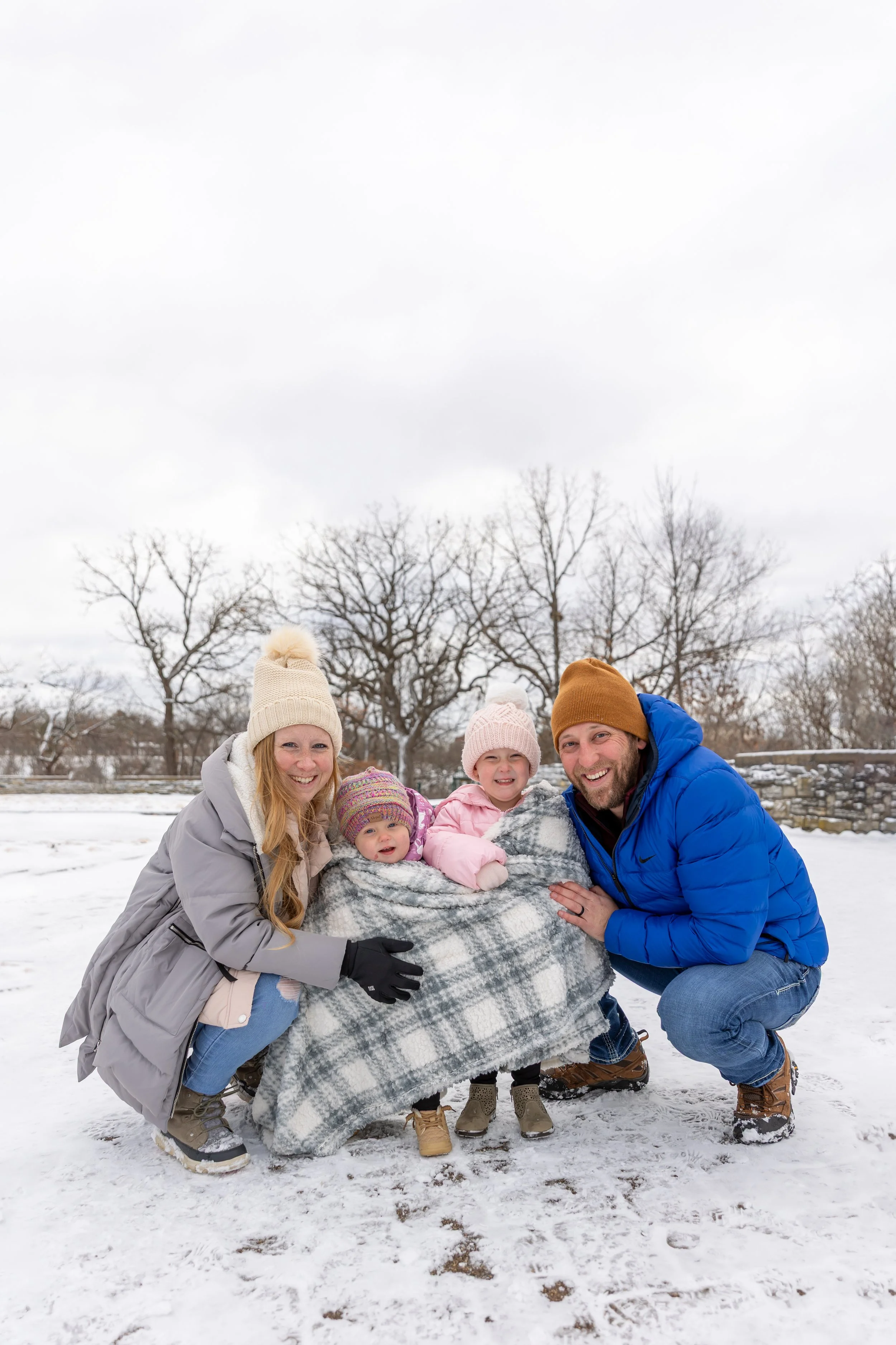 Minneapolis family photography - A mother and father squat next to their two young toddler girls as they are all bundled up in winter jackets and a blanket while smiling at the camera. It is a snowy winter scene at Minnehaha Falls in Minneapolis, MN.