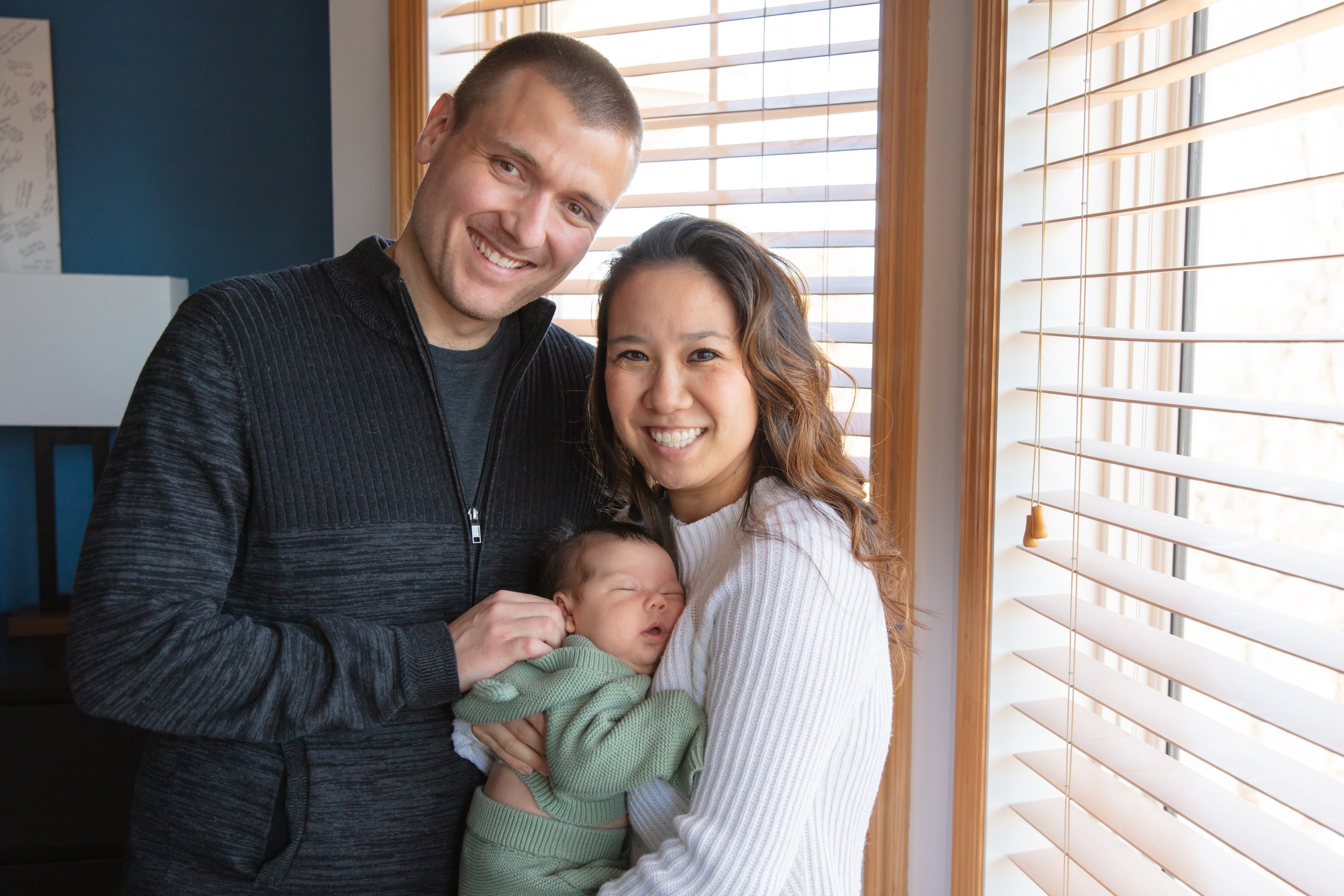 Father and mother stand together smiling at the camera while they hold their sleeping newborn baby.