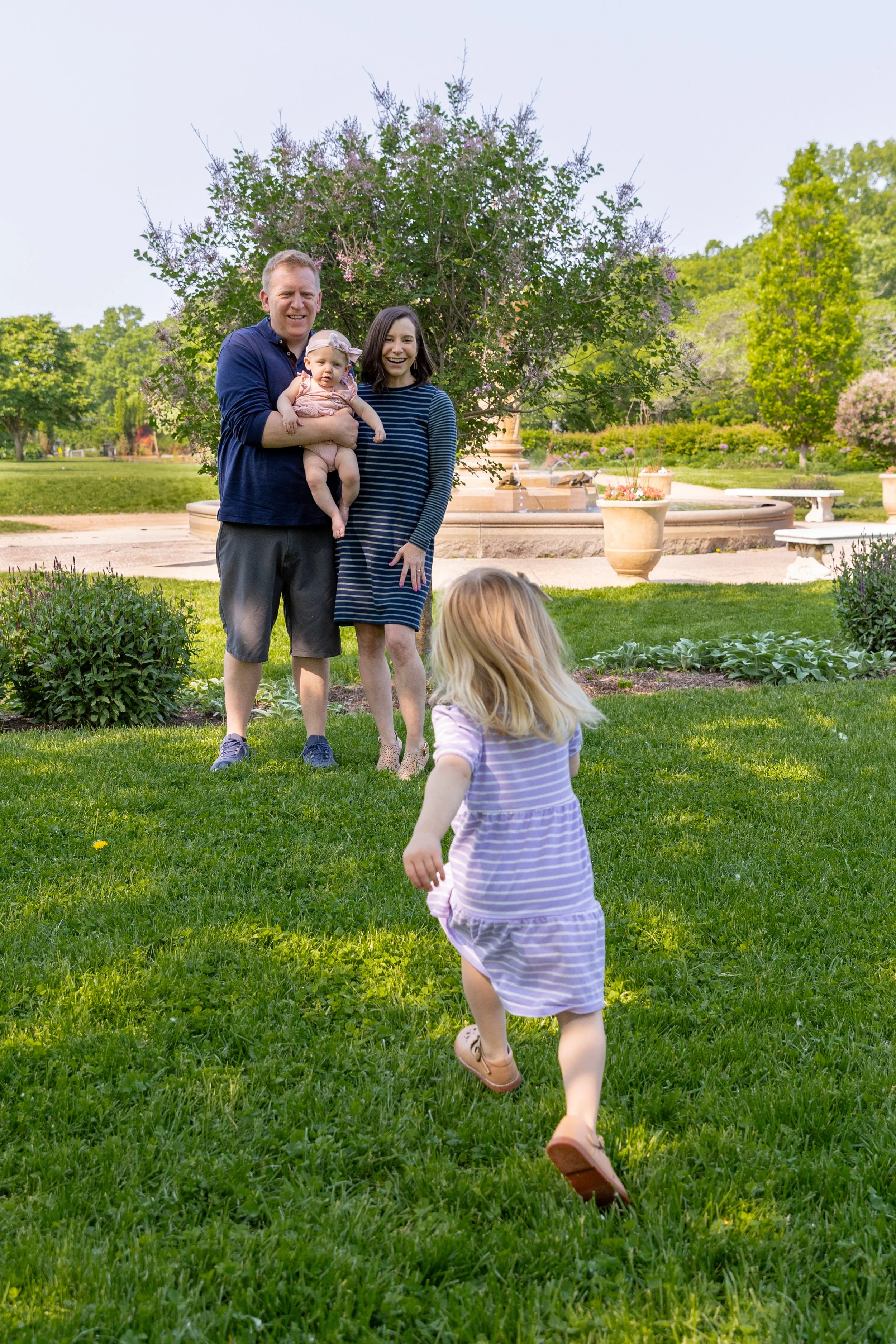 Minneapolis family photography May Mini Session- A mother and father stand in the distance with their baby girl in their arms as their older toddler runs towards them. They are at Lyndale Rose Garden in Minneapolis, MN.
