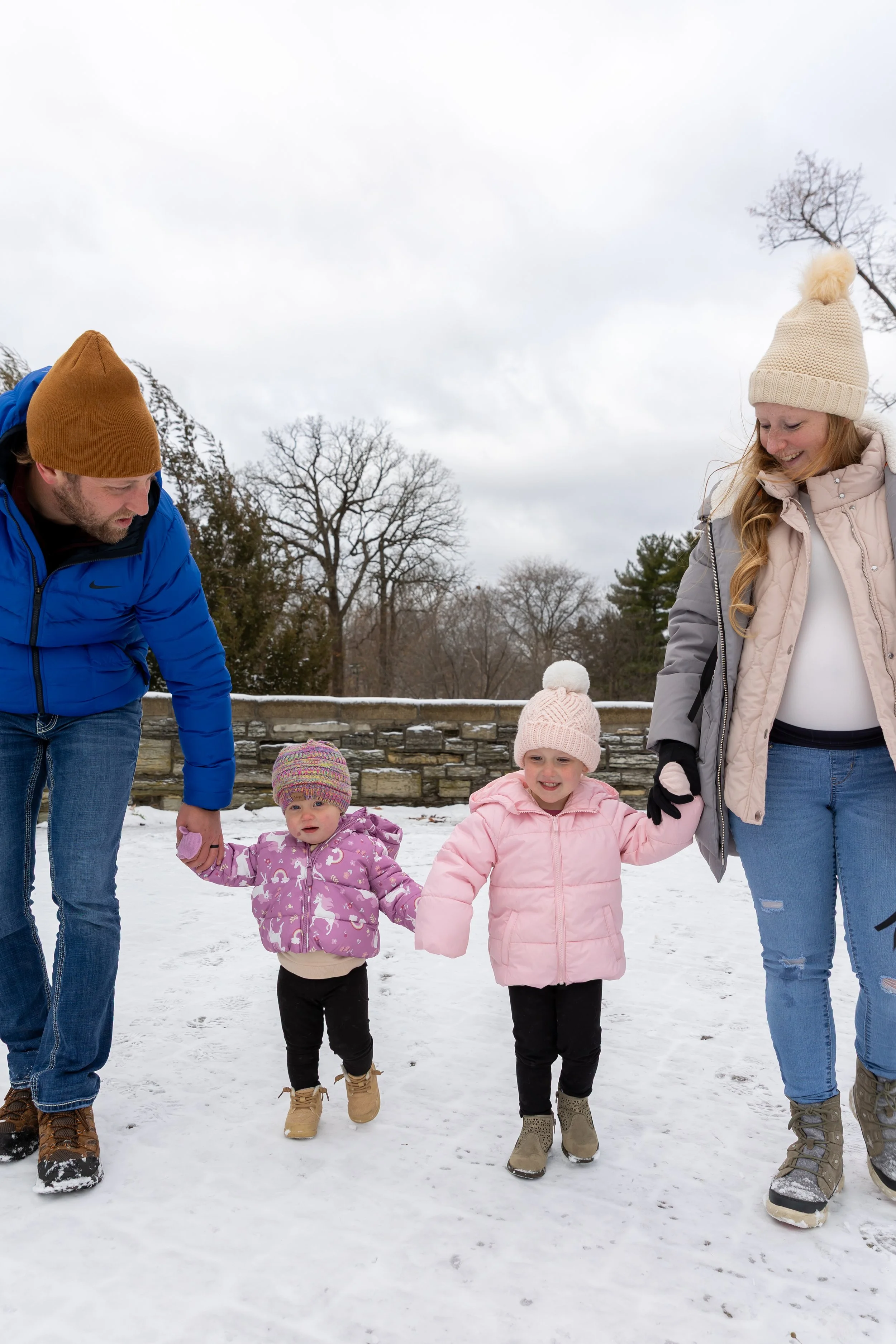 Minneapolis family photography - A mother and father hold heir two young toddler girls hands/mittens as they slowly walk towards the camera while smiling at each other. It is a snowy winter scene at Minnehaha Falls in Minneapolis, MN. 