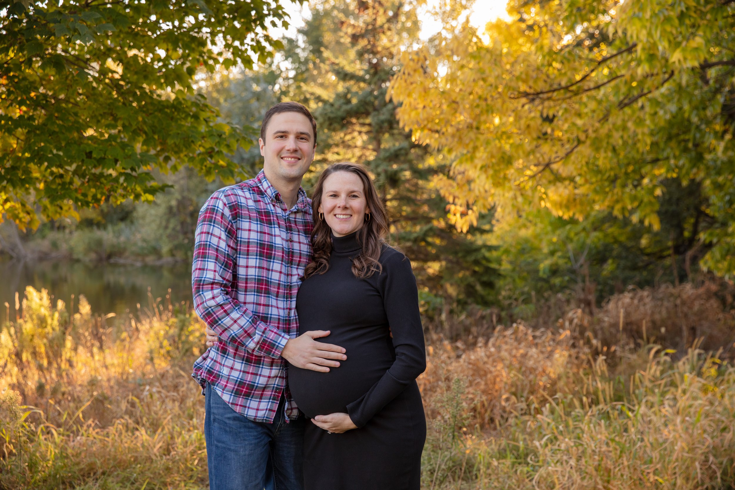 Maternity session, expecting mother and father-to-be embrace each other smiling at the camera in a Minneapolis park with fall foliage behind them.