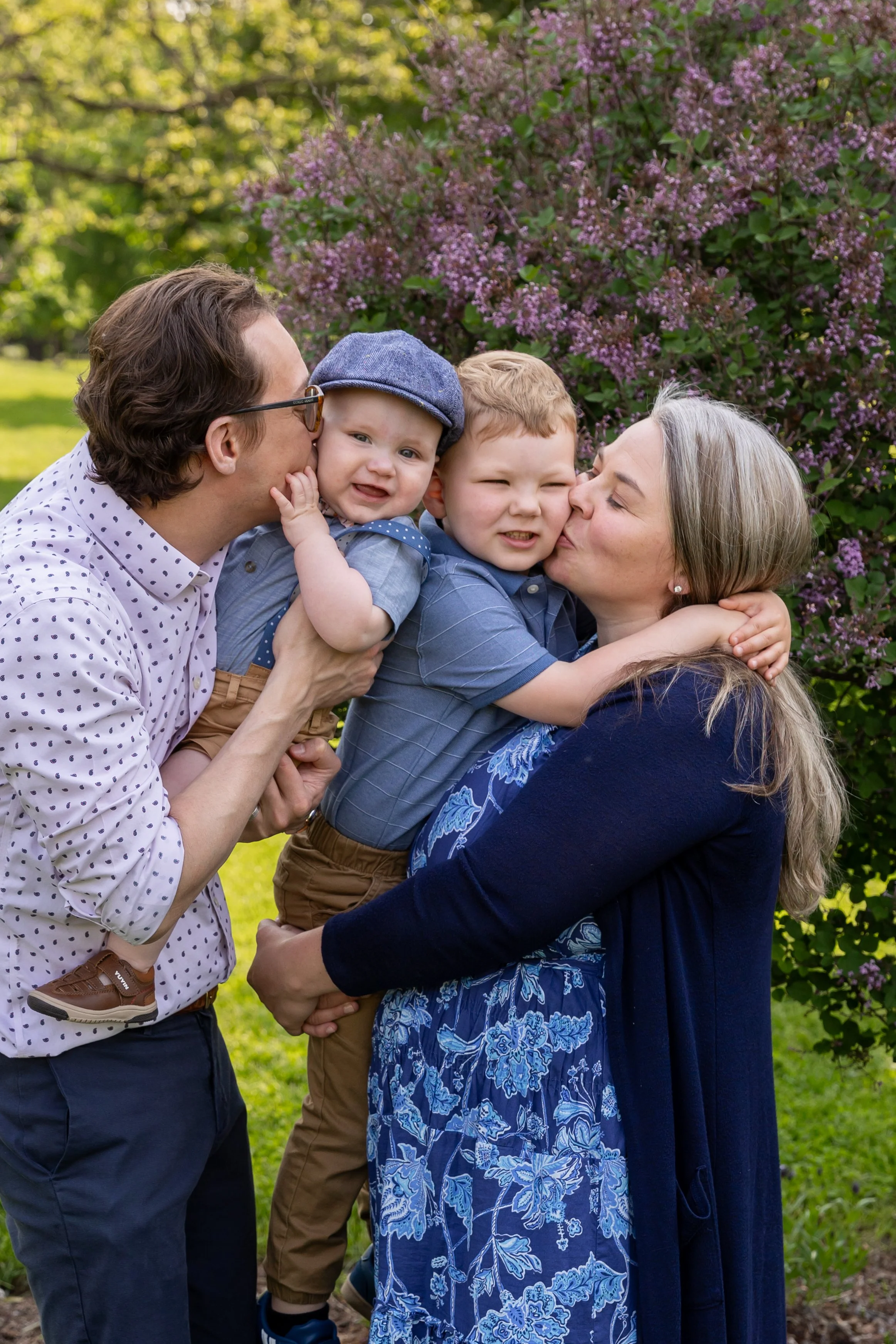 Minneapolis family photography May Mini Session- A mother and father stand together holding their toddler son and baby boy between them. The parents are kissing the cheeks of their sons. They are at Lyndale Rose Garden in Minneapolis, MN.