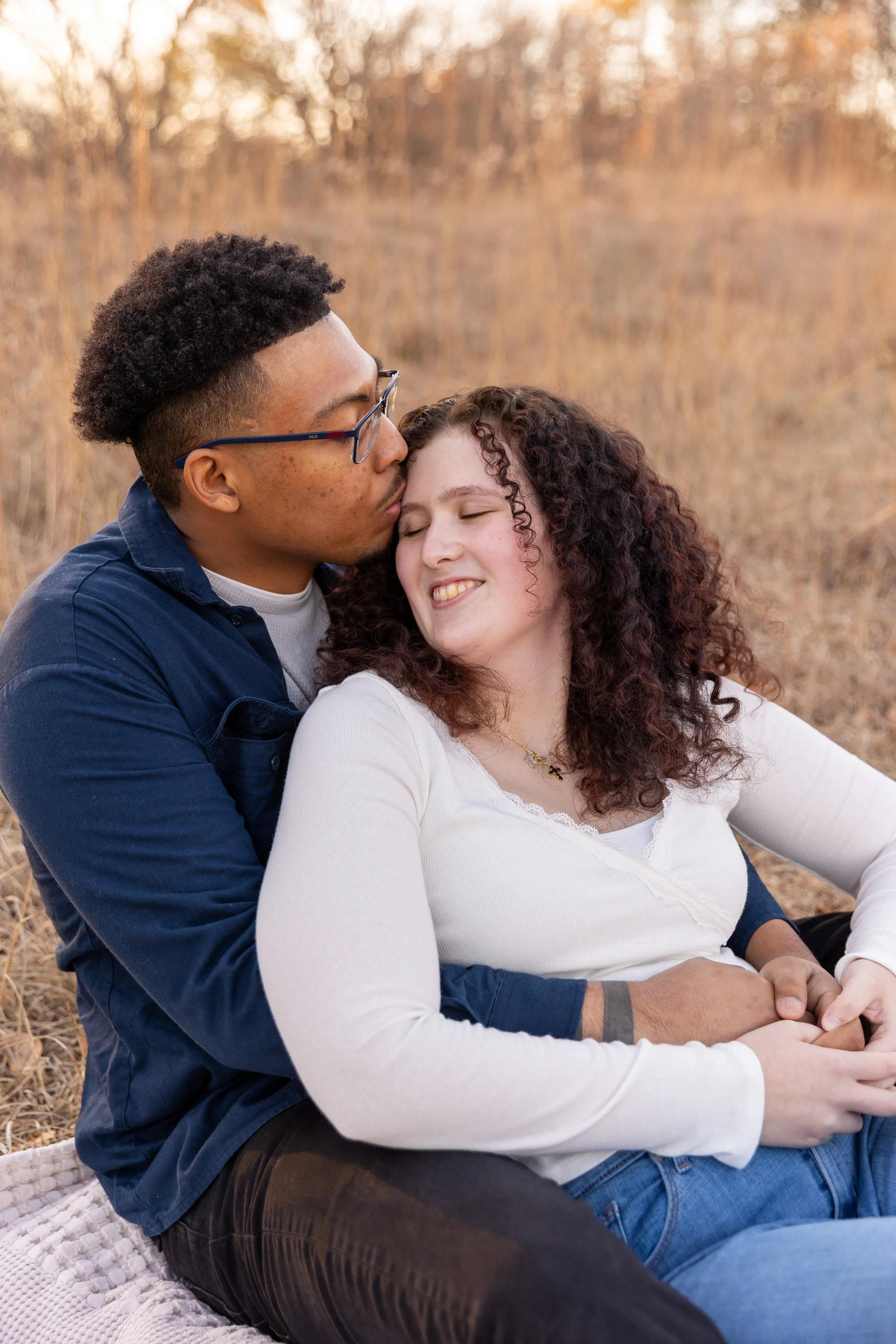 Minneapolis couples photography - A couple sits together, with the woman between the mans legs, on a blanket smiling as the man kisses the woman's forehead while at Minnehaha Falls Park in Minneapolis, MN. 