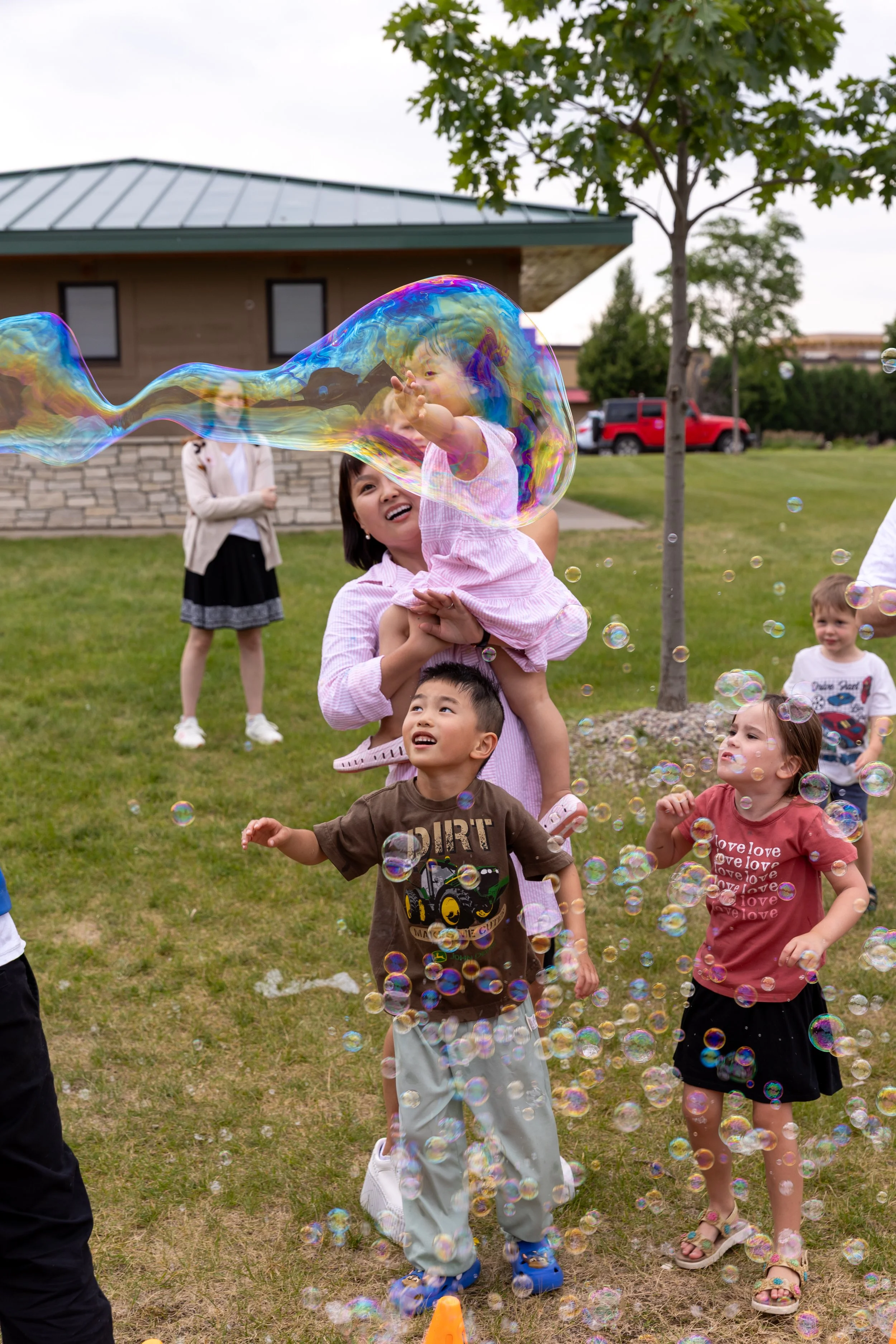 A mom in a pink dress smiles while holding her daughter up, also wearing a pink dress, in a St. Paul Park with bubbles blowing around them from a bubble machine. Other children are running and playing around them in the bubbles.