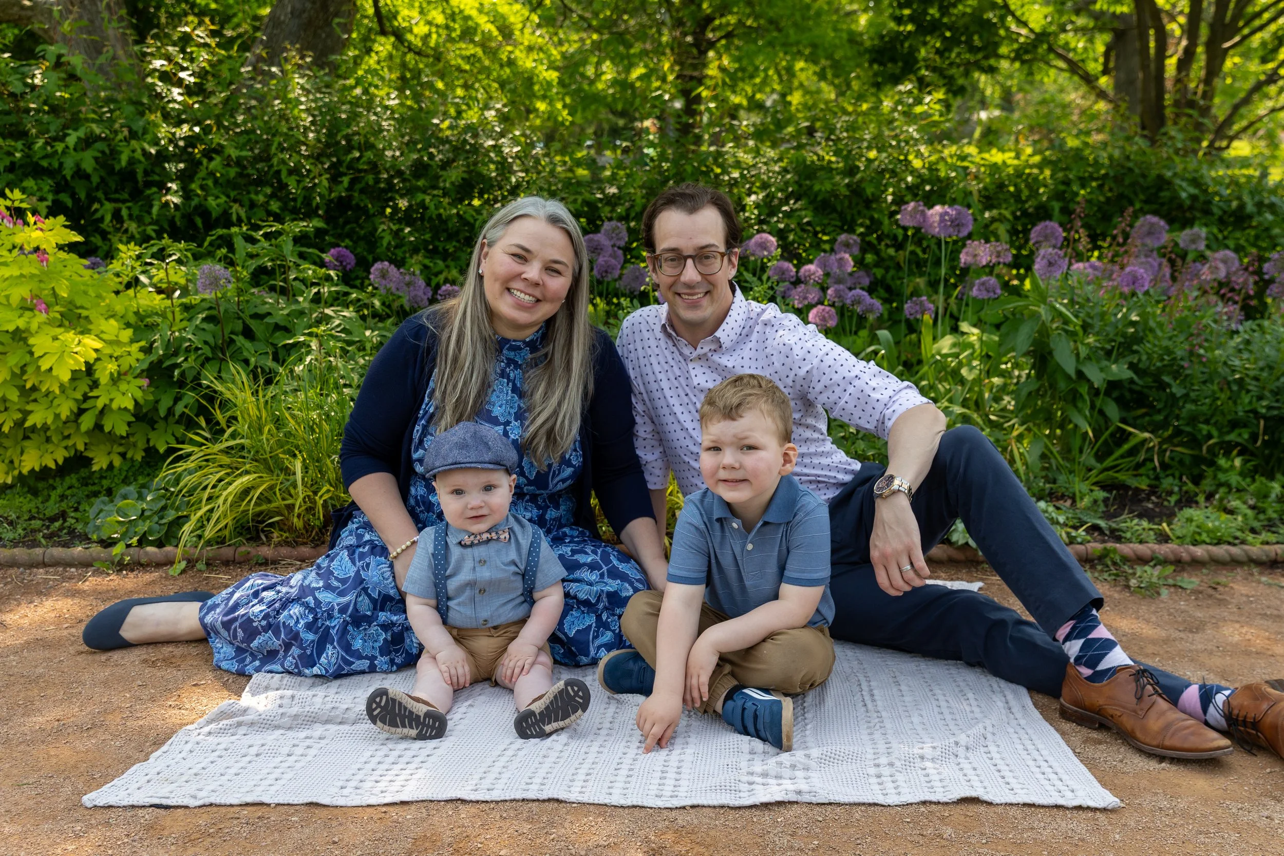 Minneapolis family photography May Mini Session- A family of four sit on a blanket together all smiling at the camera. The mother and father in the back with their toddler and baby sons in front of them. They are at Lyndale Rose Garden in Minneapolis