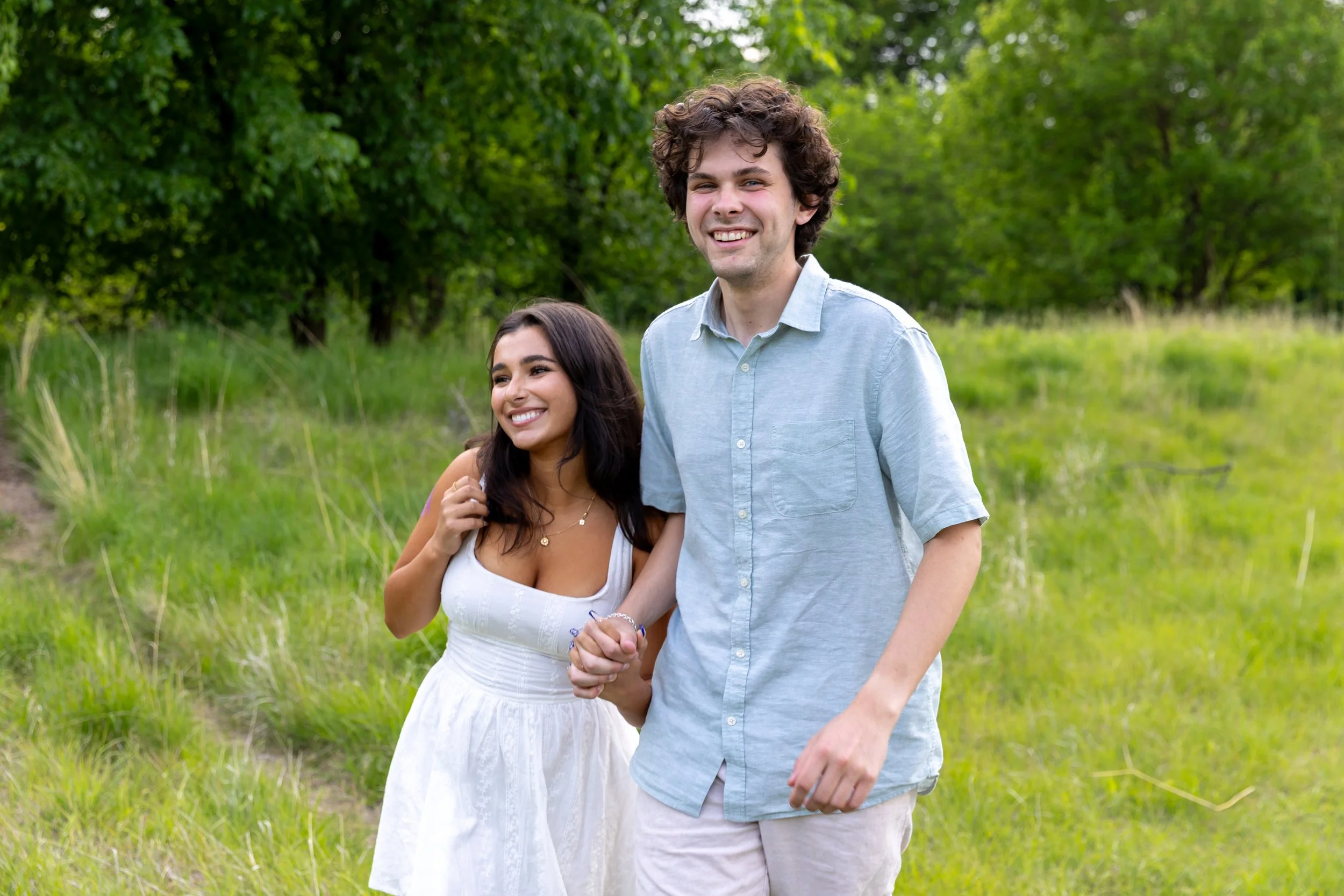 Minneapolis couples photography - A couple holds hands while walking towards the camera in a grassy field smiling while at Minnehaha Falls Park in Minneapolis, MN. 