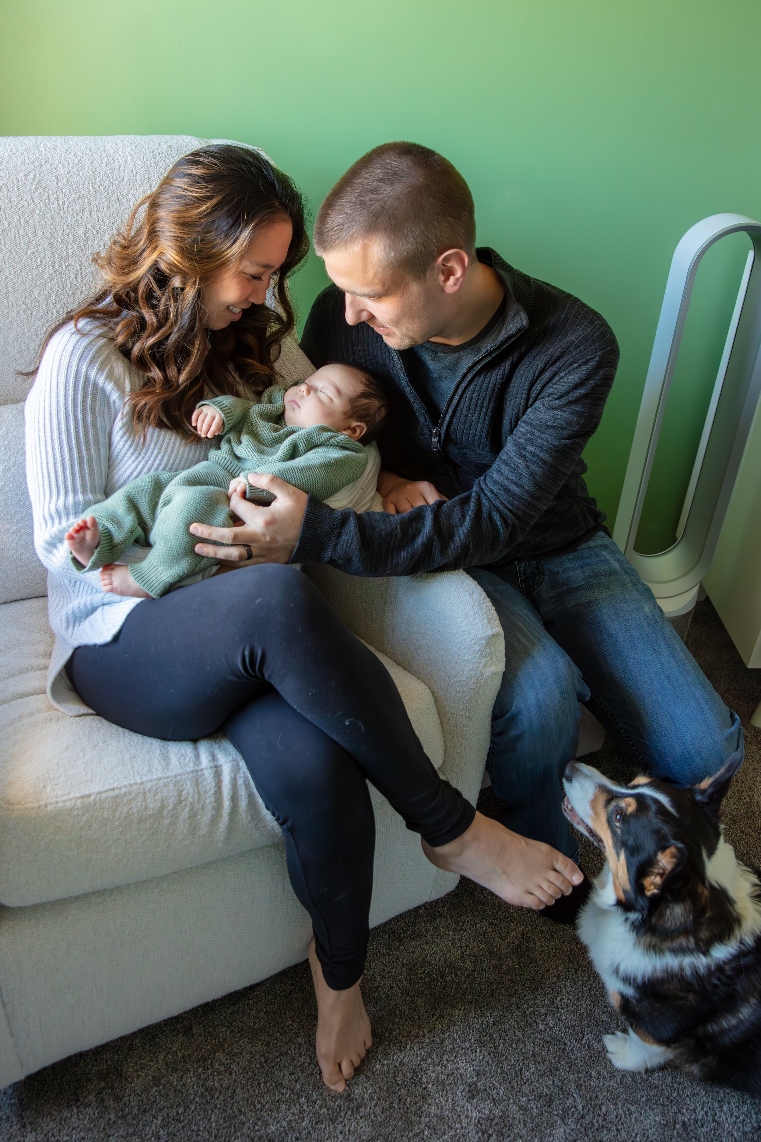 Newborn session, mother and father smile looking down with their heads together while holding their sleeping baby. Their corgi dog is sitting on the ground in front of them looking up at the family.
