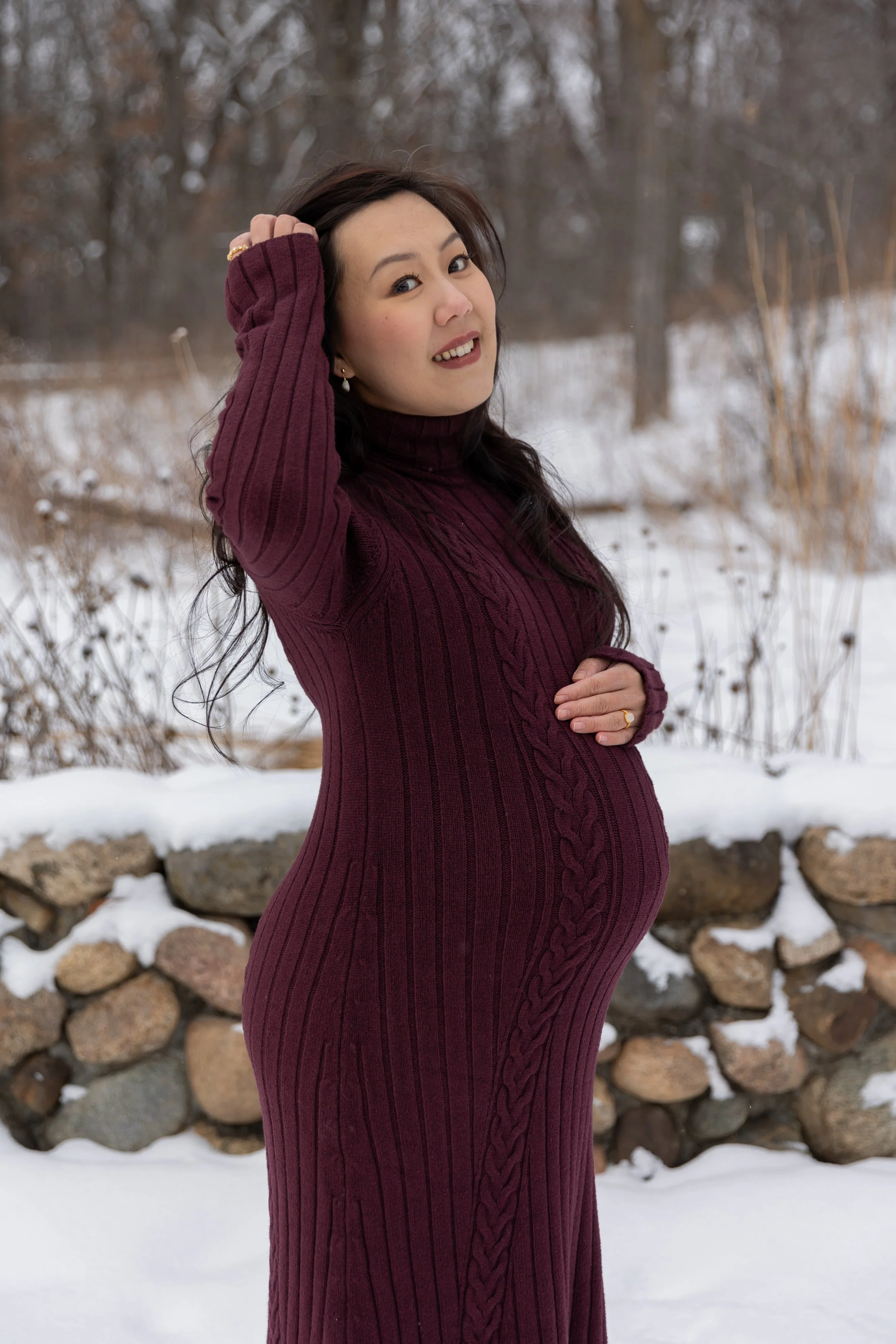 Minneapolis Maternity session, expecting mother cradles her baby bump while brushing her hair back and smiling at the camera at Minnetonka Mills Park in Hopkins, MN in the winter.