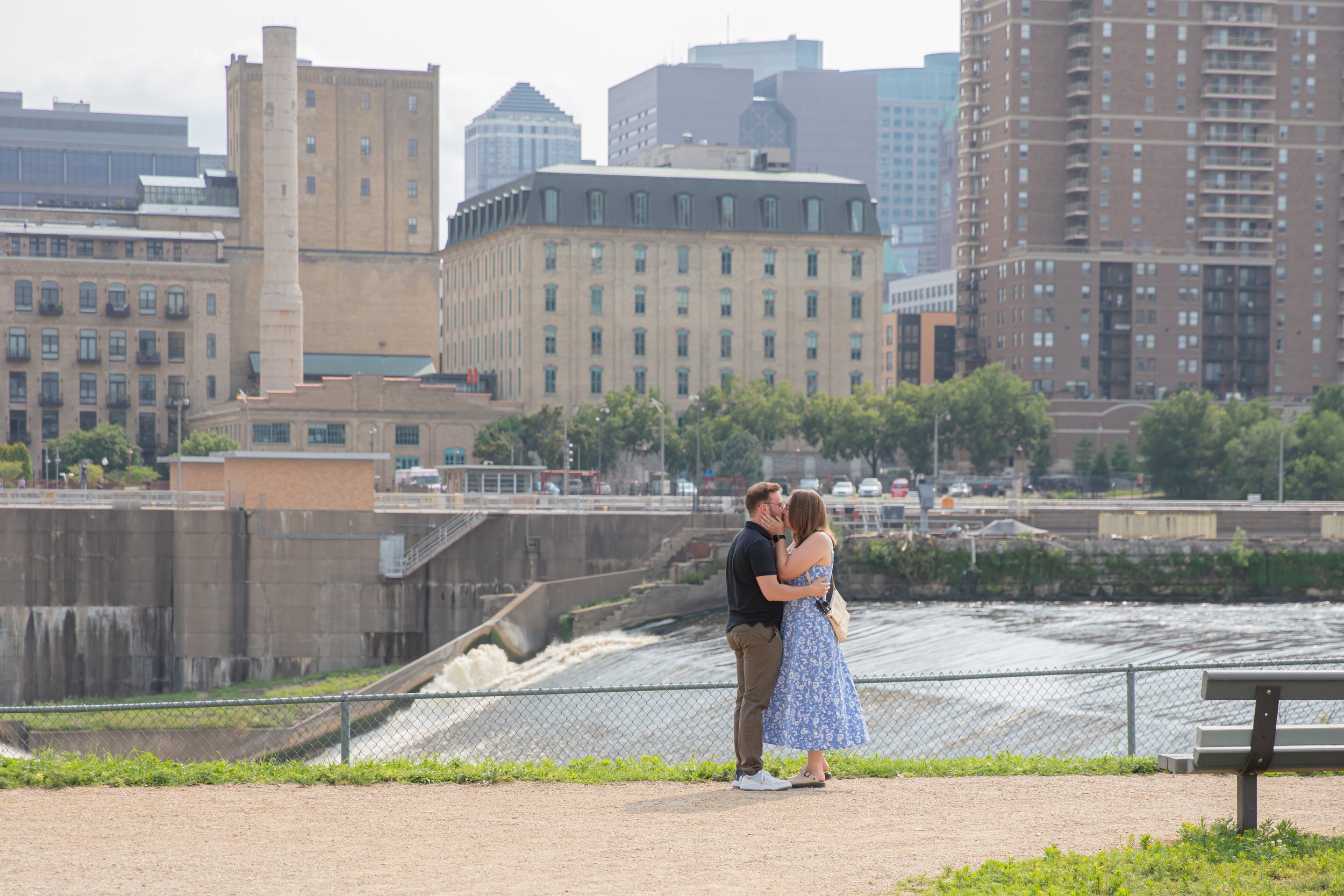 A couple, a man and woman, embraces each other kissing on a park path right after he proposed to her in front of the falls and skyline at Water Power Park on St. Anthony Main.
