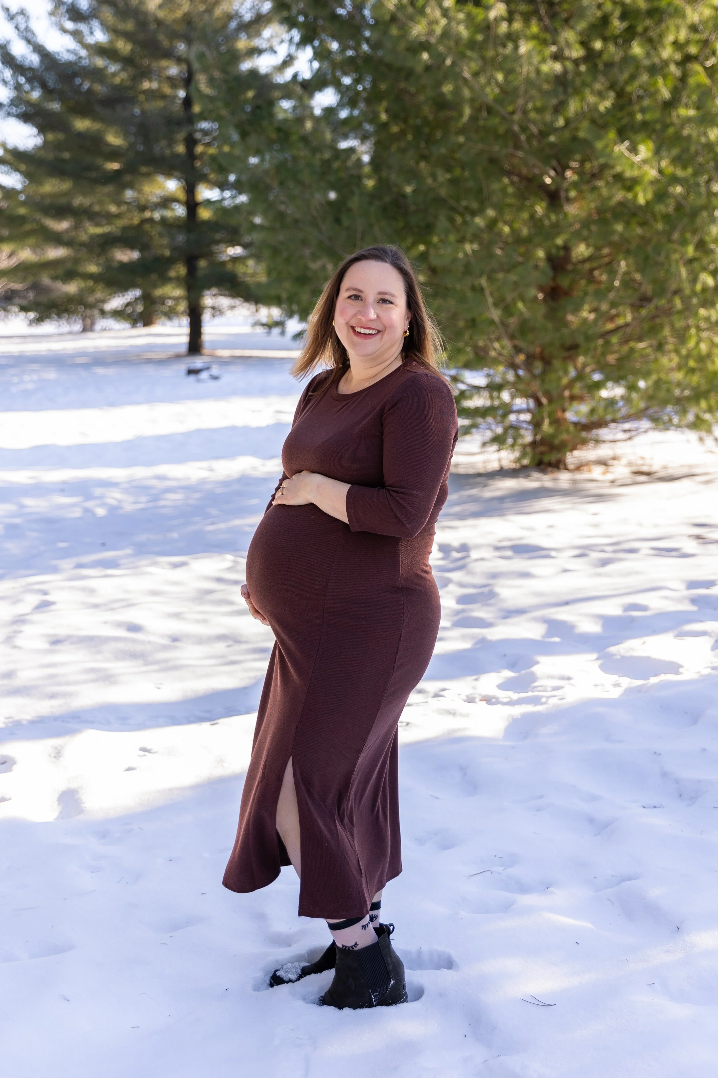 Minneapolis Maternity session, expecting mother cradles her baby bump smiling at the camera at Arneson Acres Park in Edina, MN in the winter.