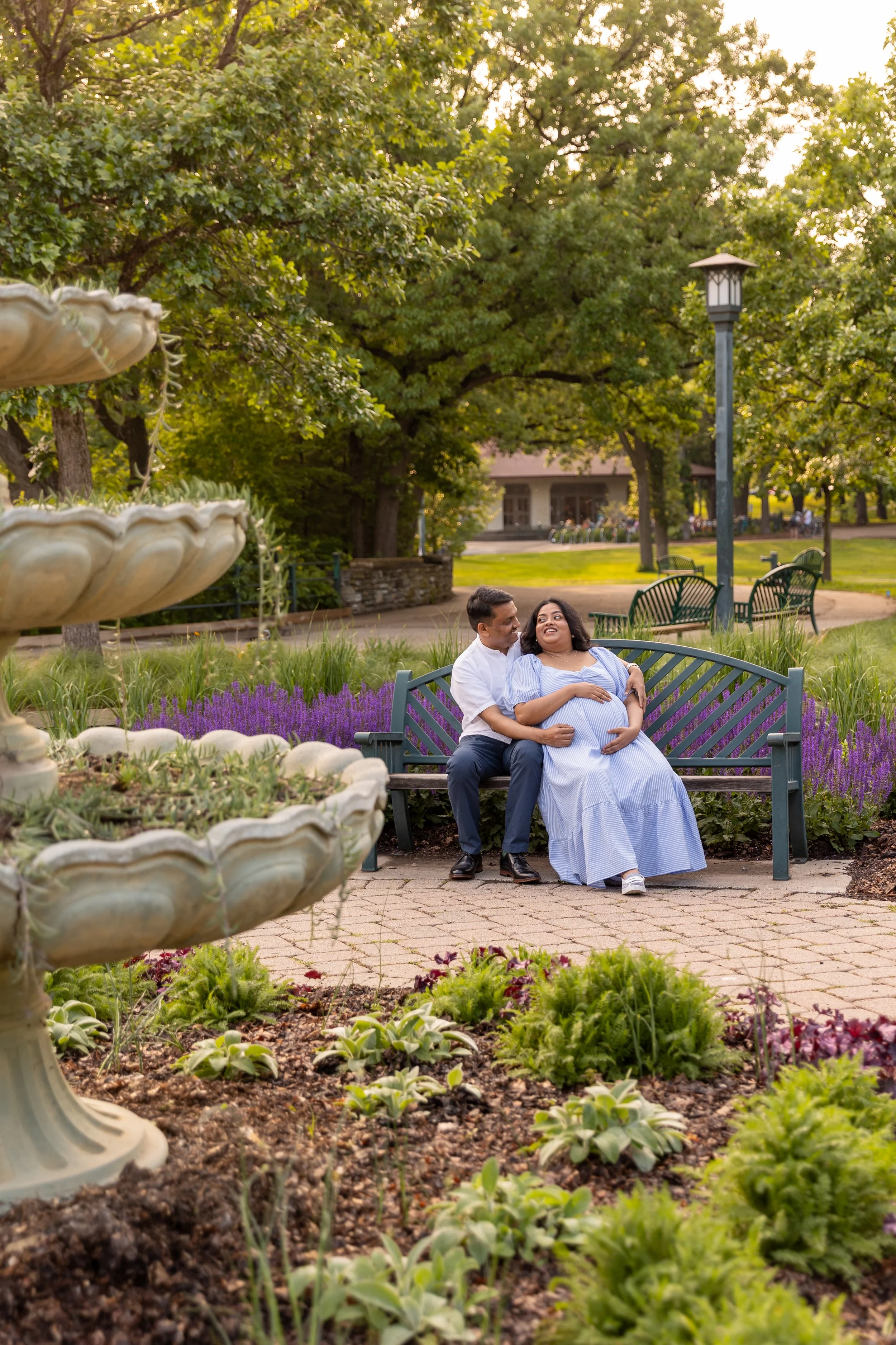 Maternity session - father and mother sit on a bench together while gazing lovingly at each other at Minnehaha Falls Park while holding the baby bump