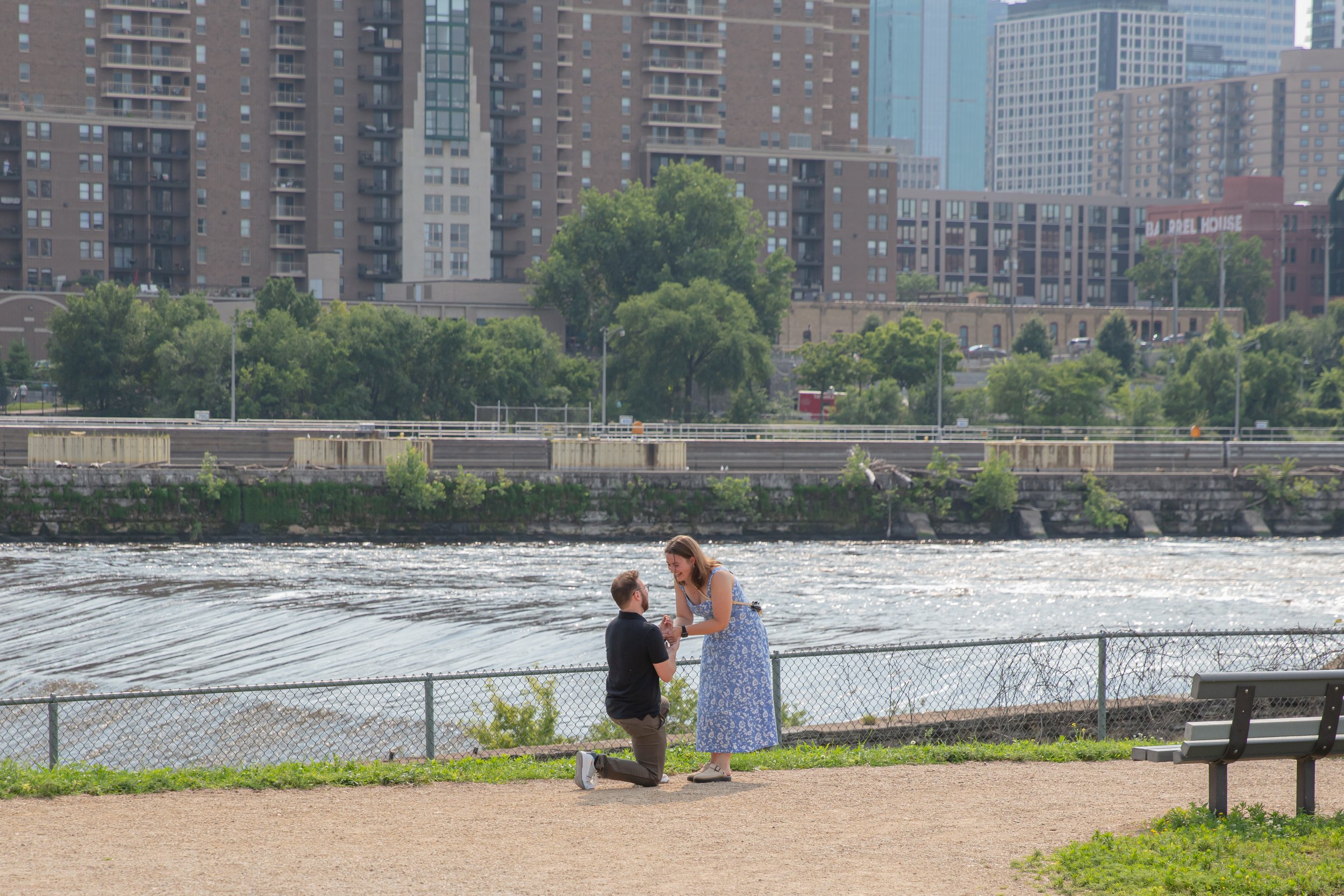 A man kneels in front of a woman on a park path proposing to her in front of the falls and skyline at Water Power Park on St. Anthony Main.
