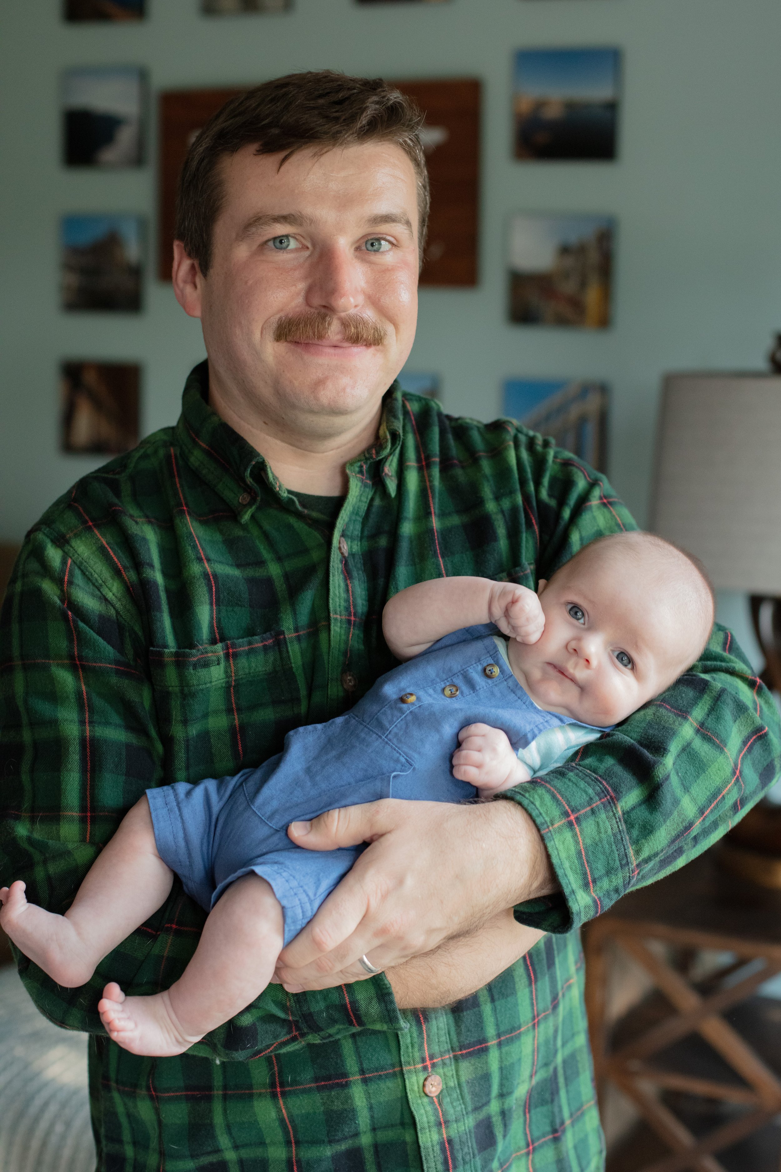 Dad stands in his living room while holding his baby and smiling at the camera.