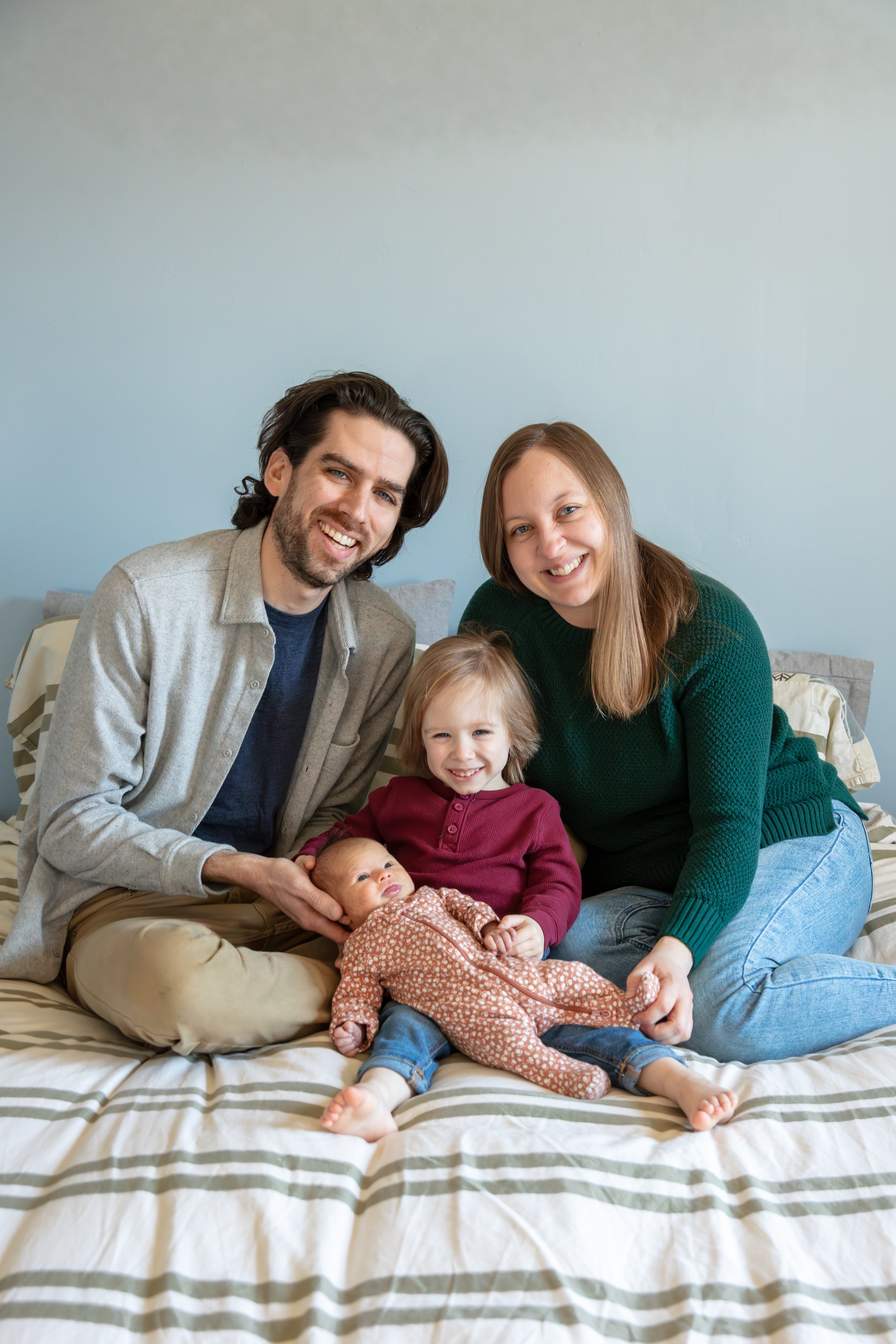 A father and mother sit on their bed with their toddler between them as he holds his newborn sister, all smiling at the camera.