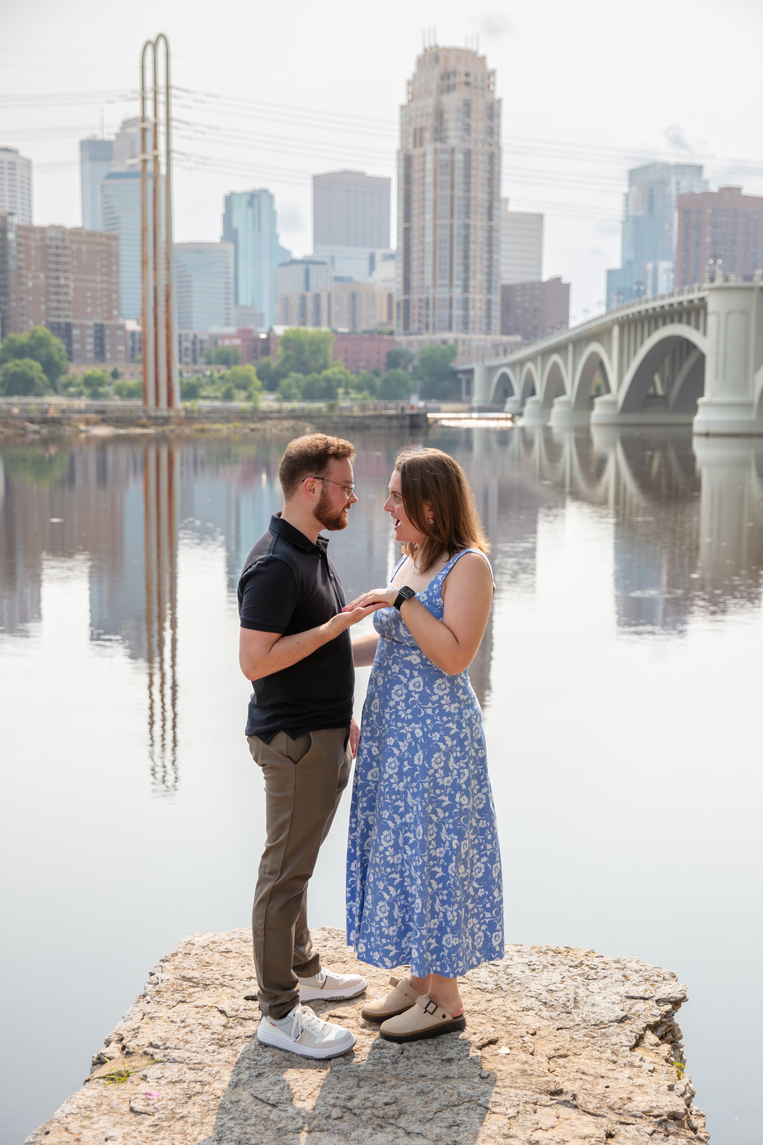 A couple, a man and woman, smile and look at the woman's ring hand between them while on standing a rock after he proposed to her in front of the falls and skyline at Water Power Park on St. Anthony Main.