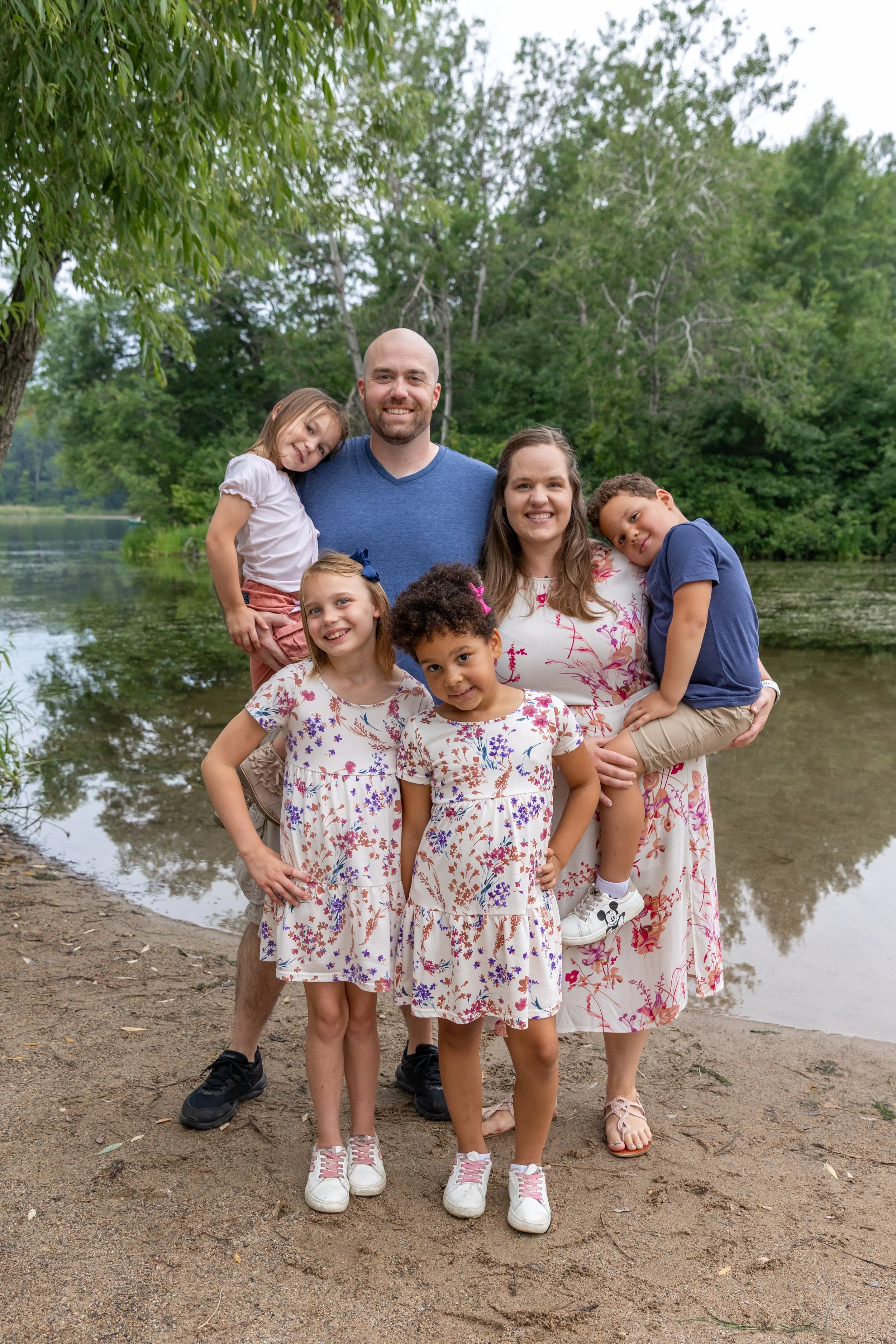 Minneapolis Family Photography - A blended family of a mom, dad and four kids stand together smiling at the camera on a beach with the lake in the background at Lebanon Hills Regional Park in Eagan, MN. 