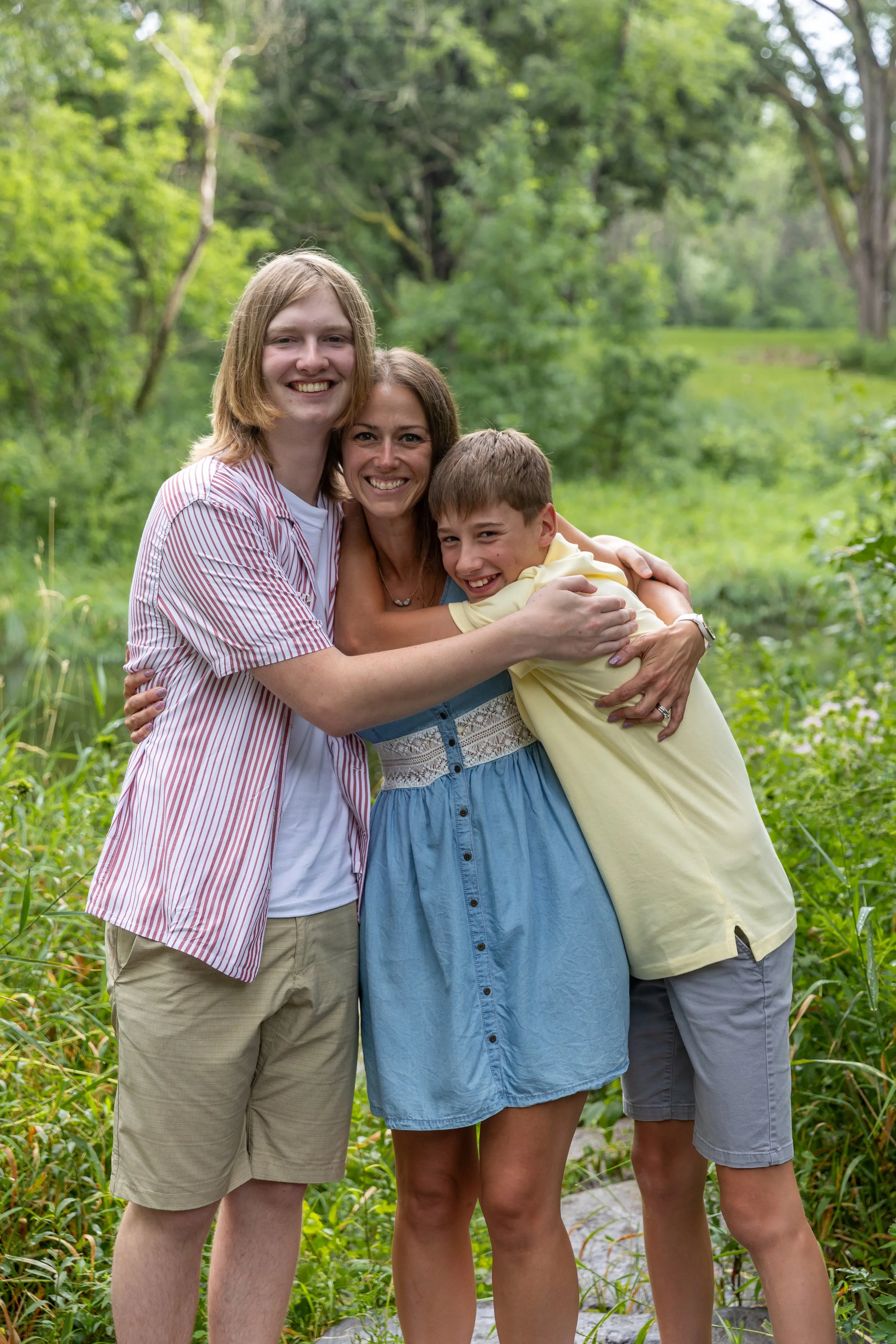 A mother stands in the middle of her two teenage sons who both embrace her at Minnetonka Mills Park in Hopkins, MN while all smiling at the camera. 