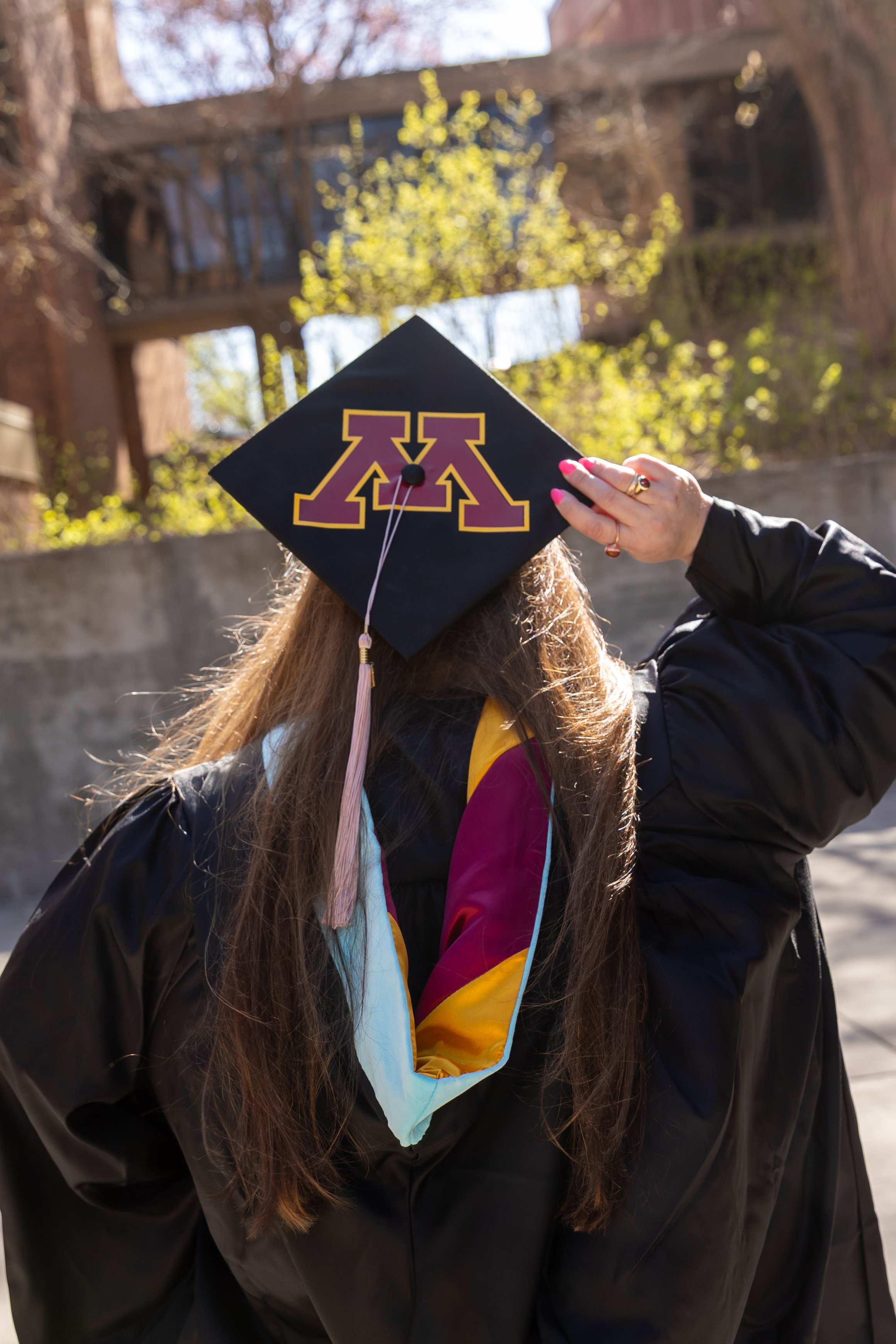 A University of Minnesota masters graduate holds her graduation cap dressed in her graduation robe on the U of M campus while facing away from the camera with the focus on the M logo of the university on the cap.