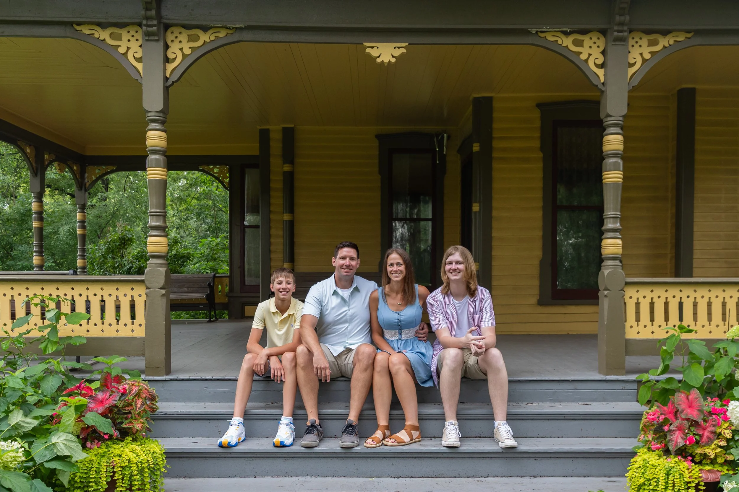 A family of four - a father and mother sit in the middle of their two teenage sons - sits on the front steps of a yellow and green Victorian style home at Minnetonka Mills Park in Hopkins, MN smiling at the camera. 
