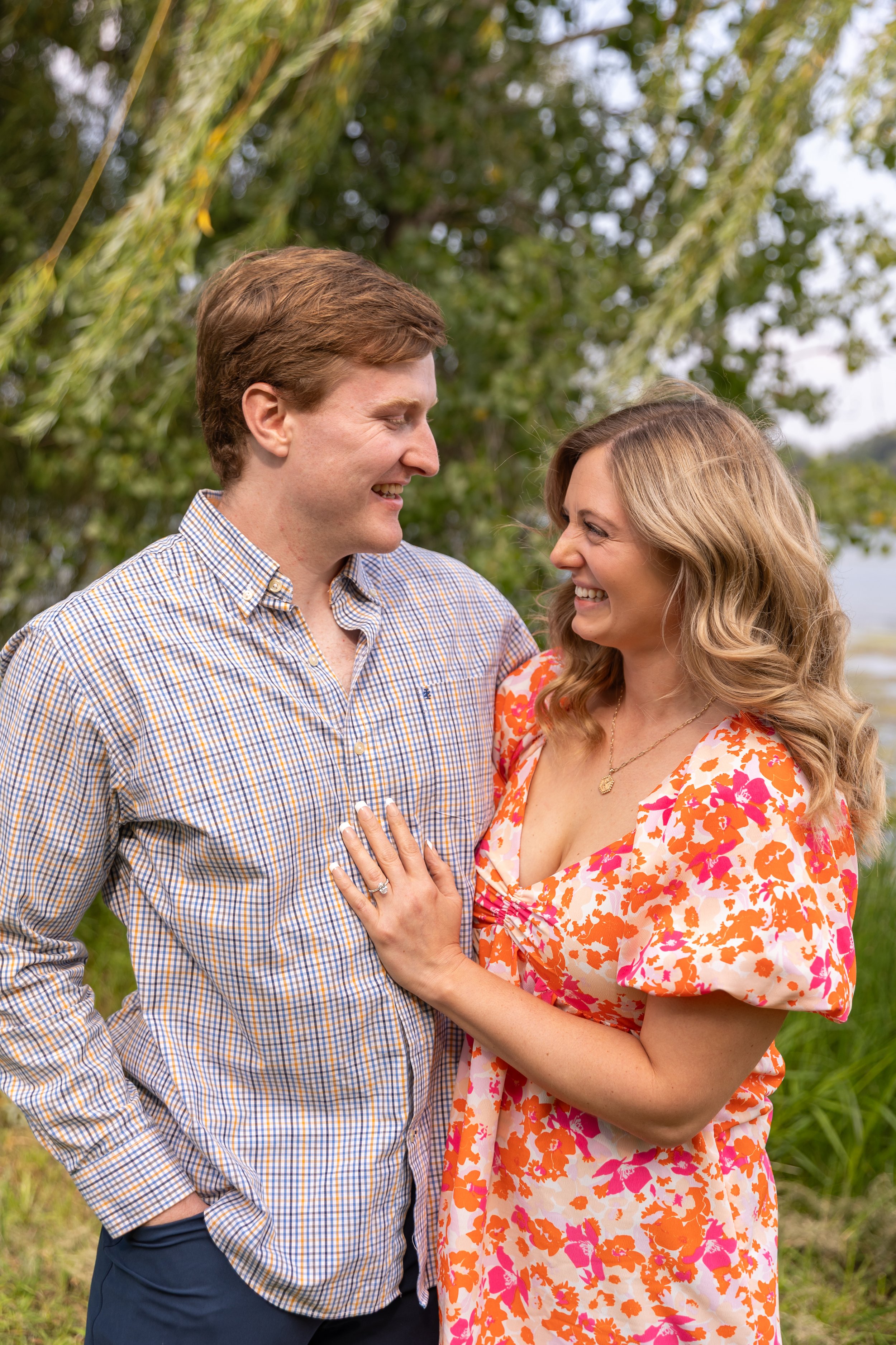 Minneapolis surprise proposal photography - A man and woman stand cuddled together smiling at each other at Lake Harriet. She has her left handon his chest as she shows her new engagement ring.