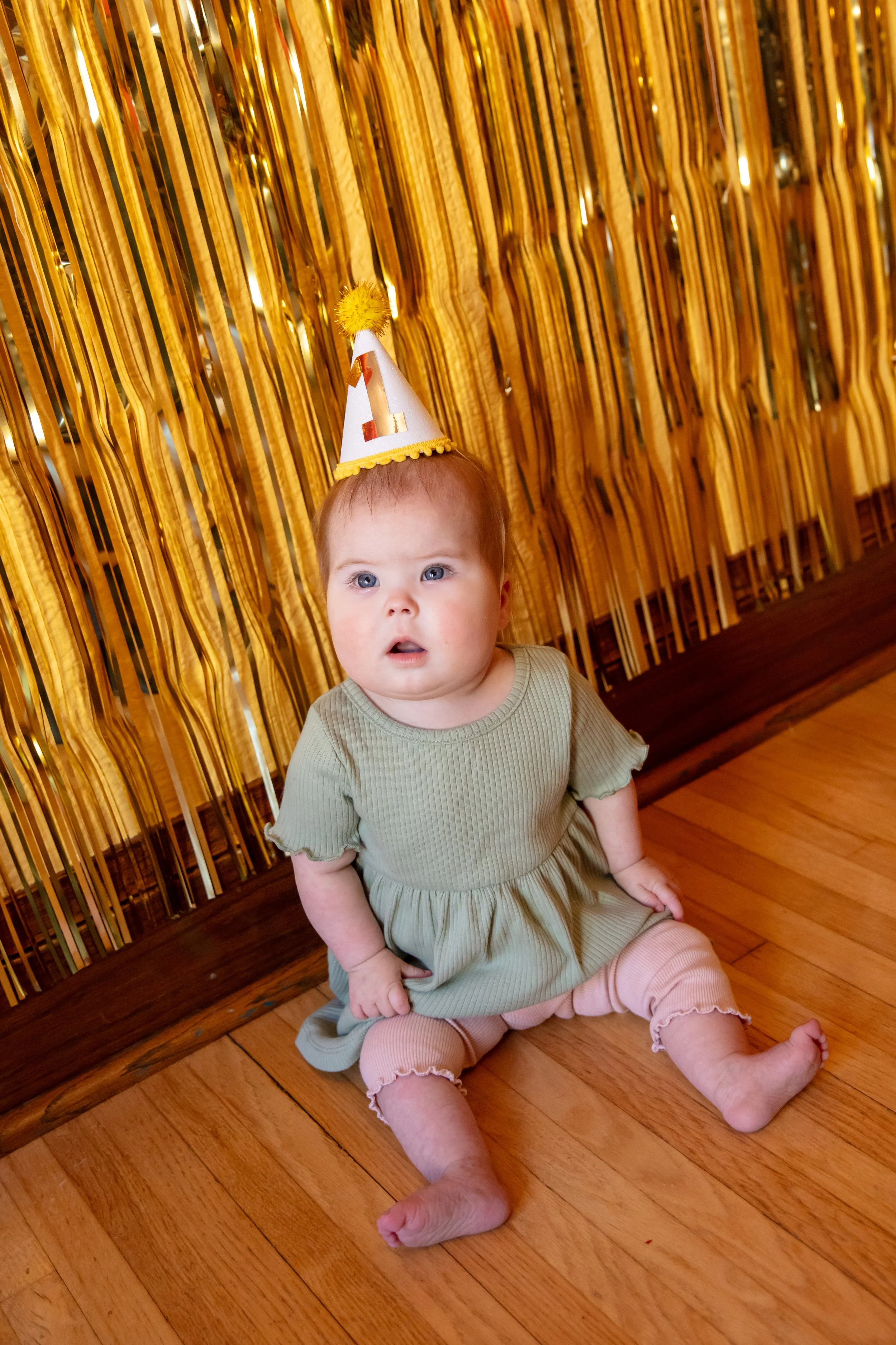 Minneapolis Cake Smash photographer - photo of a one year old baby is sitting on the floor looking up at the camera and is sitting in front of birthday streamers.  She is wearing a green dress and a birthday hat with the number 1 on it.