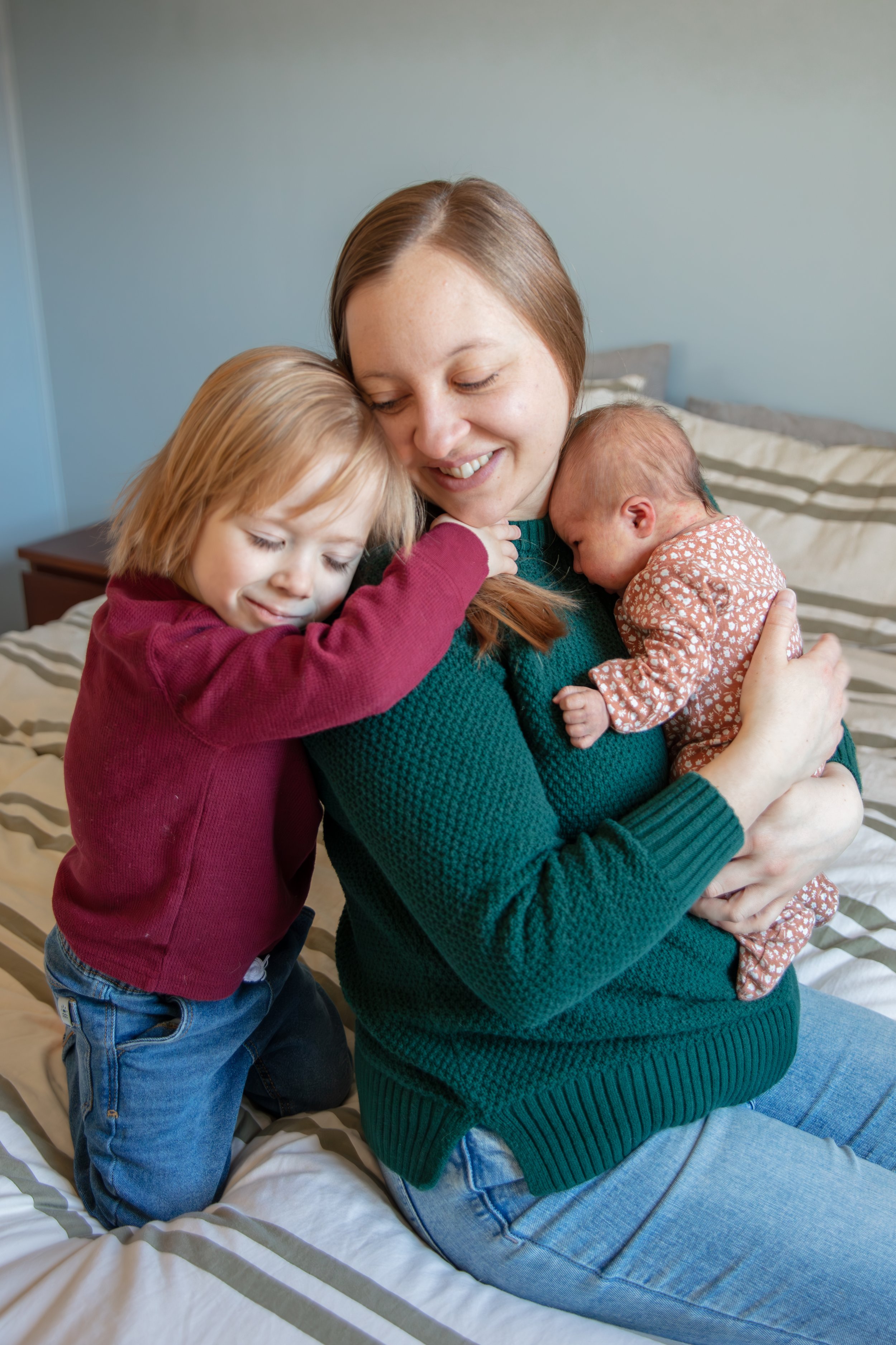 A mother holds her newborn on her chest as her toddler hugs her from behind.