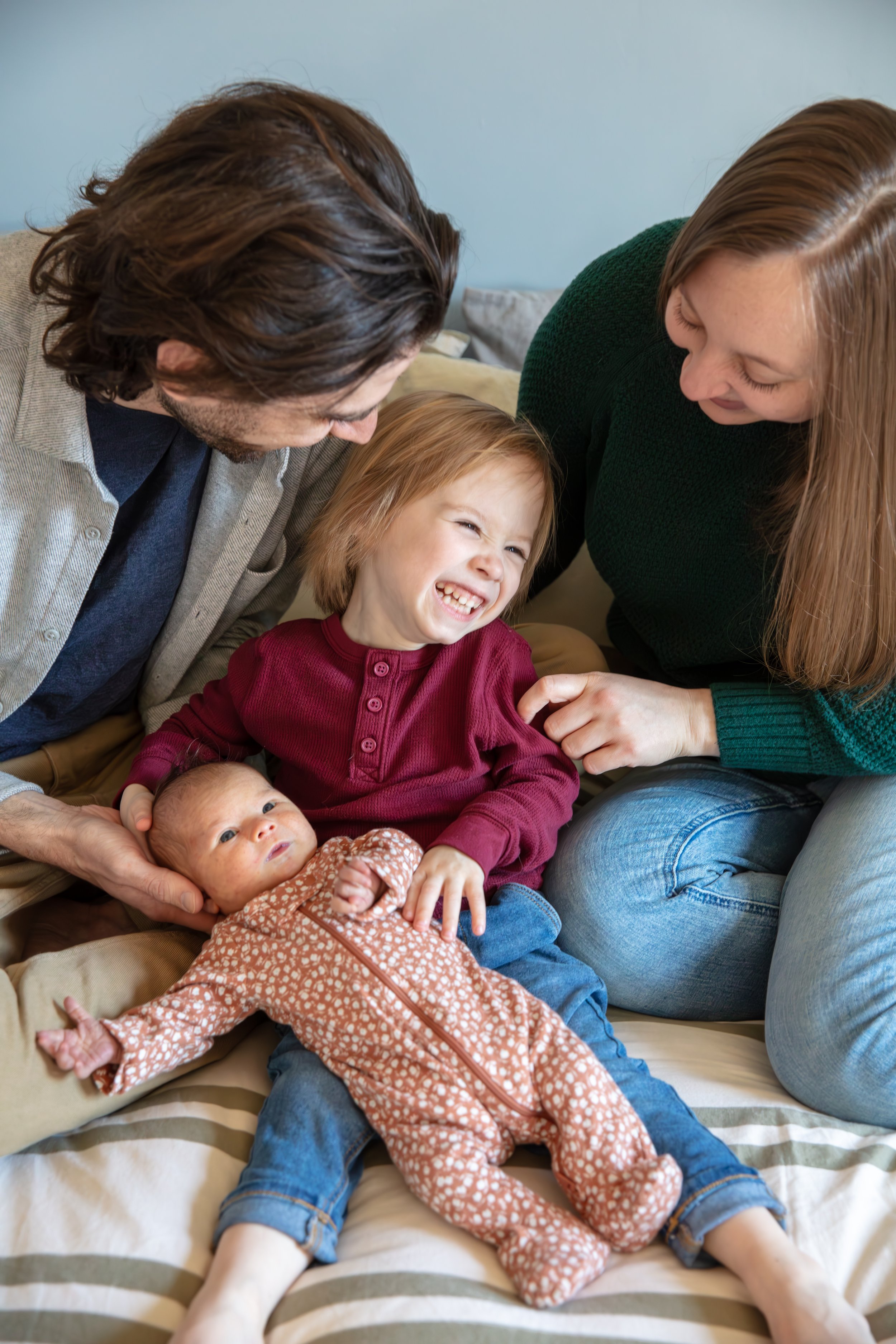 A father and mother sit on their bed tickling their toddler between them as he holds his newborn sister.