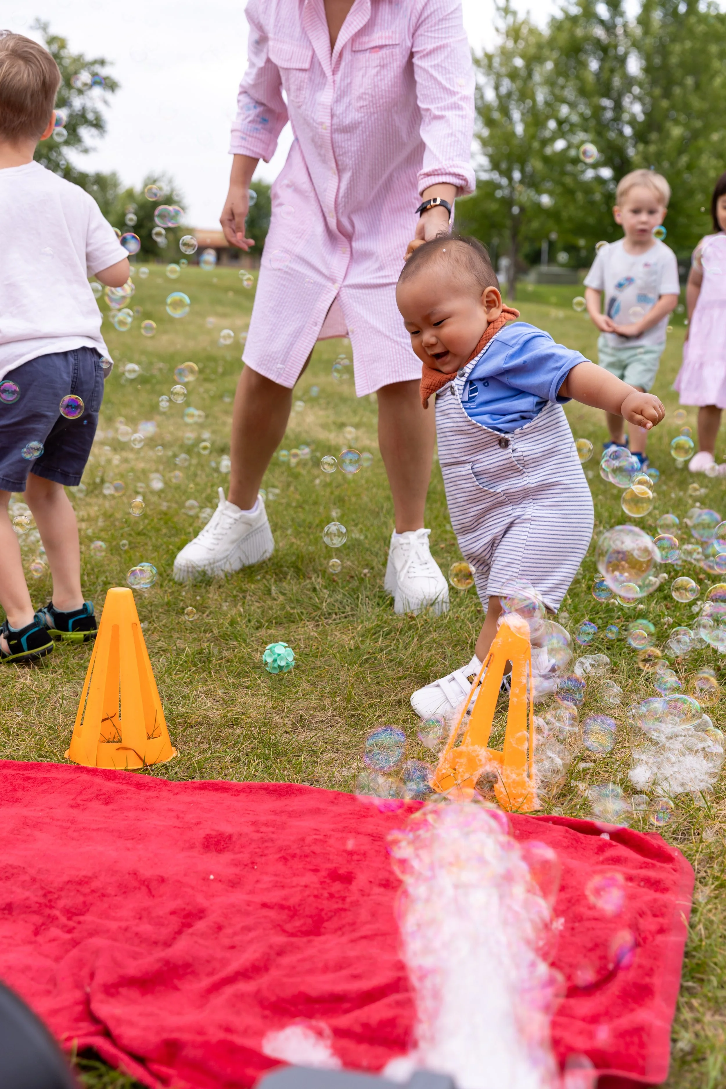 A one year old in white and blue striped overalls dances around bubbles blowing from a bubble machine in a St. Paul park his mom in a pink dress holding his hand. Other children are running and playing around them in the bubbles.
