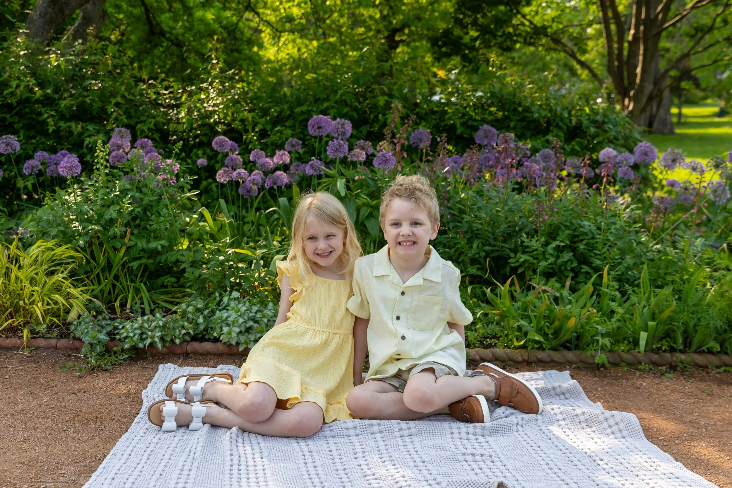 Minneapolis family photography May Mini Session- Two twin children, a sister and brother, sit on a blanket together smiling at the camera. They are at Lyndale Rose Garden in Minneapolis, MN.