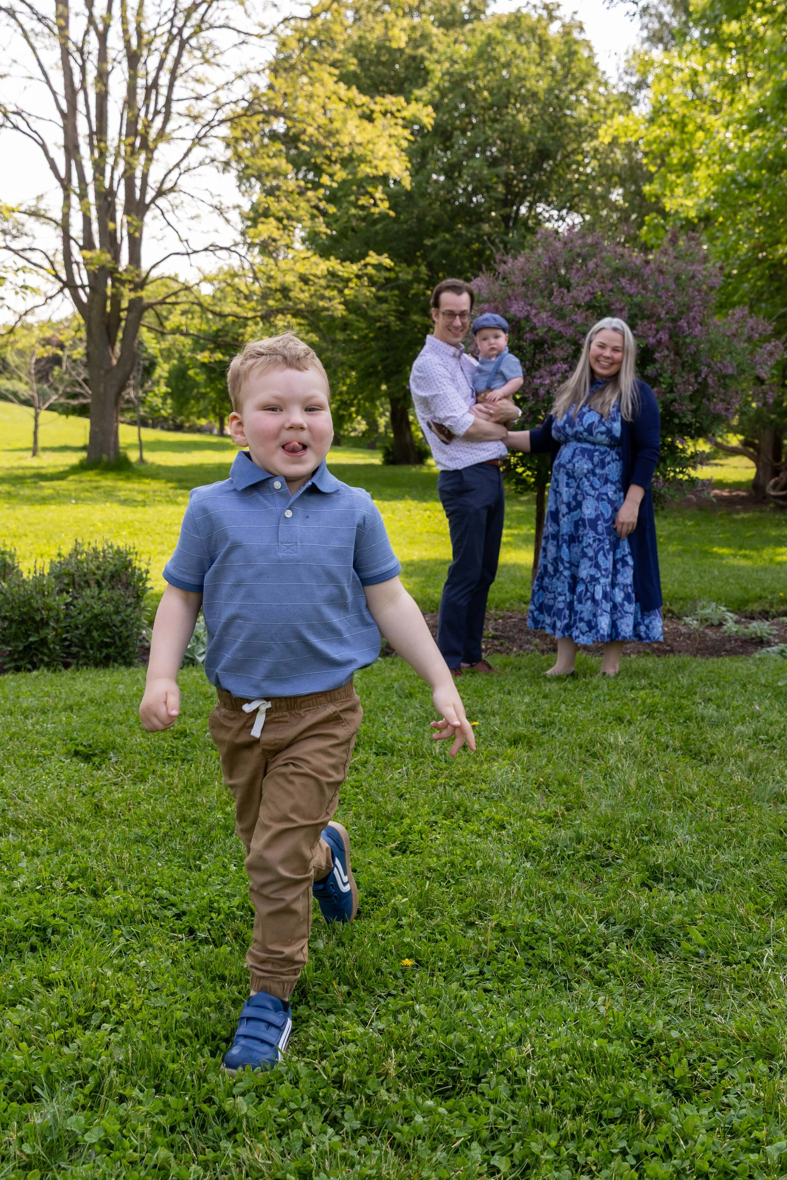 Minneapolis family photography May Mini Session- A mother and father stand in the distance with their baby boy in their arms as their older toddler runs towards the camera smiling. They are at Lyndale Rose Garden in Minneapolis, MN.