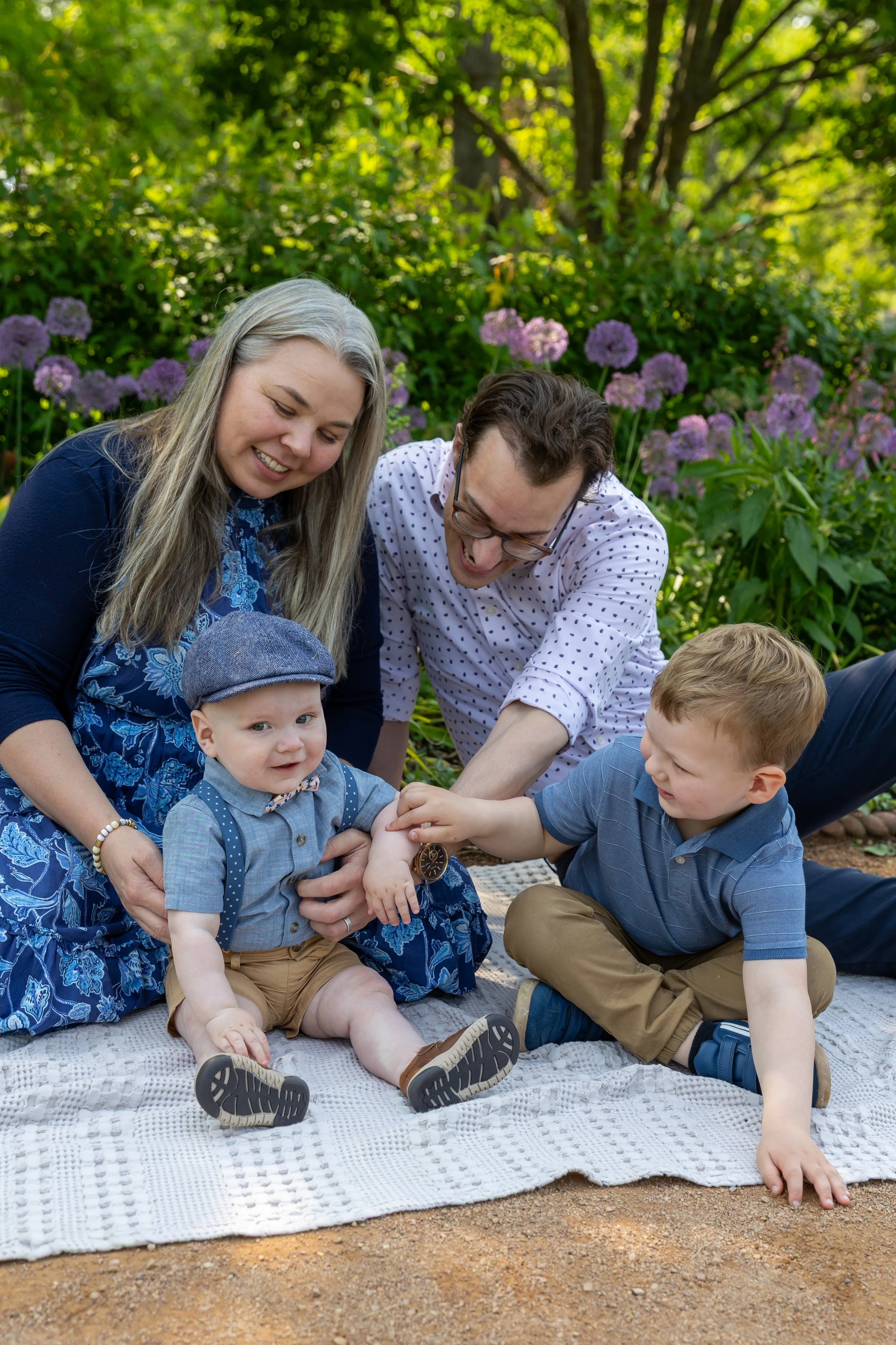 Minneapolis family photography May Mini Session- A family of four sit on a blanket together with the parents tickling their sons all laughing together. The mother and father in the back with their toddler and baby sons in front of them. They are at L
