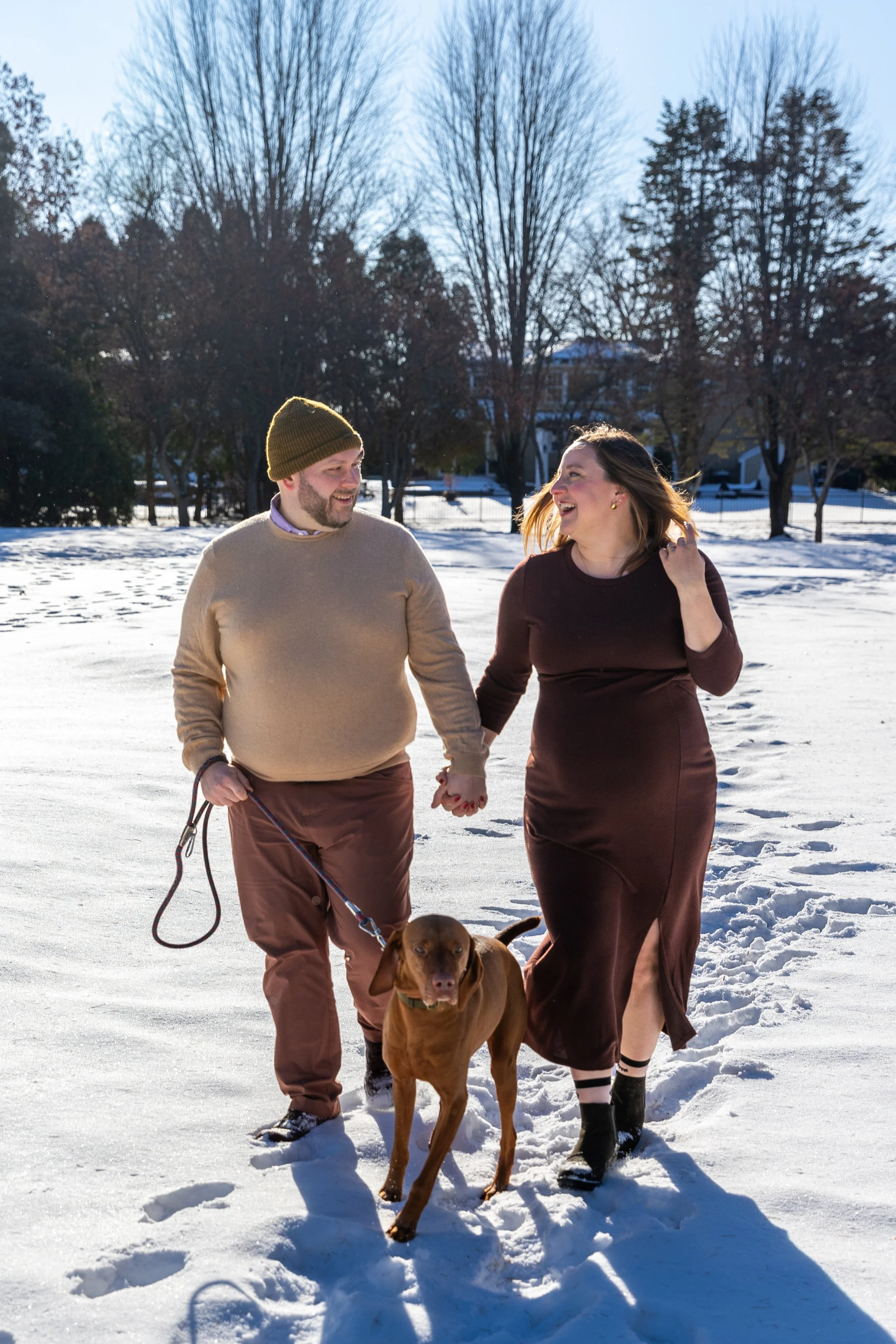 Minneapolis Maternity session, an expecting mother and father walk hand in hand smiling at each other with their dog walking in front of them at Arneson Acres Park in Edina, MN in the winter.