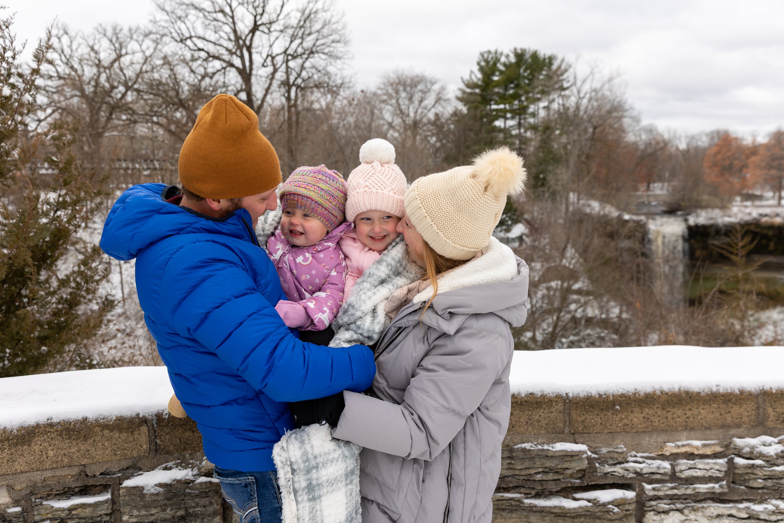 Minneapolis family photography - A mother and father are standing with a waterfall in the background as they hold their two young toddler girls as they are all bundled up in winter jackets and a blanket while smiling at each other. It is a snowy wint